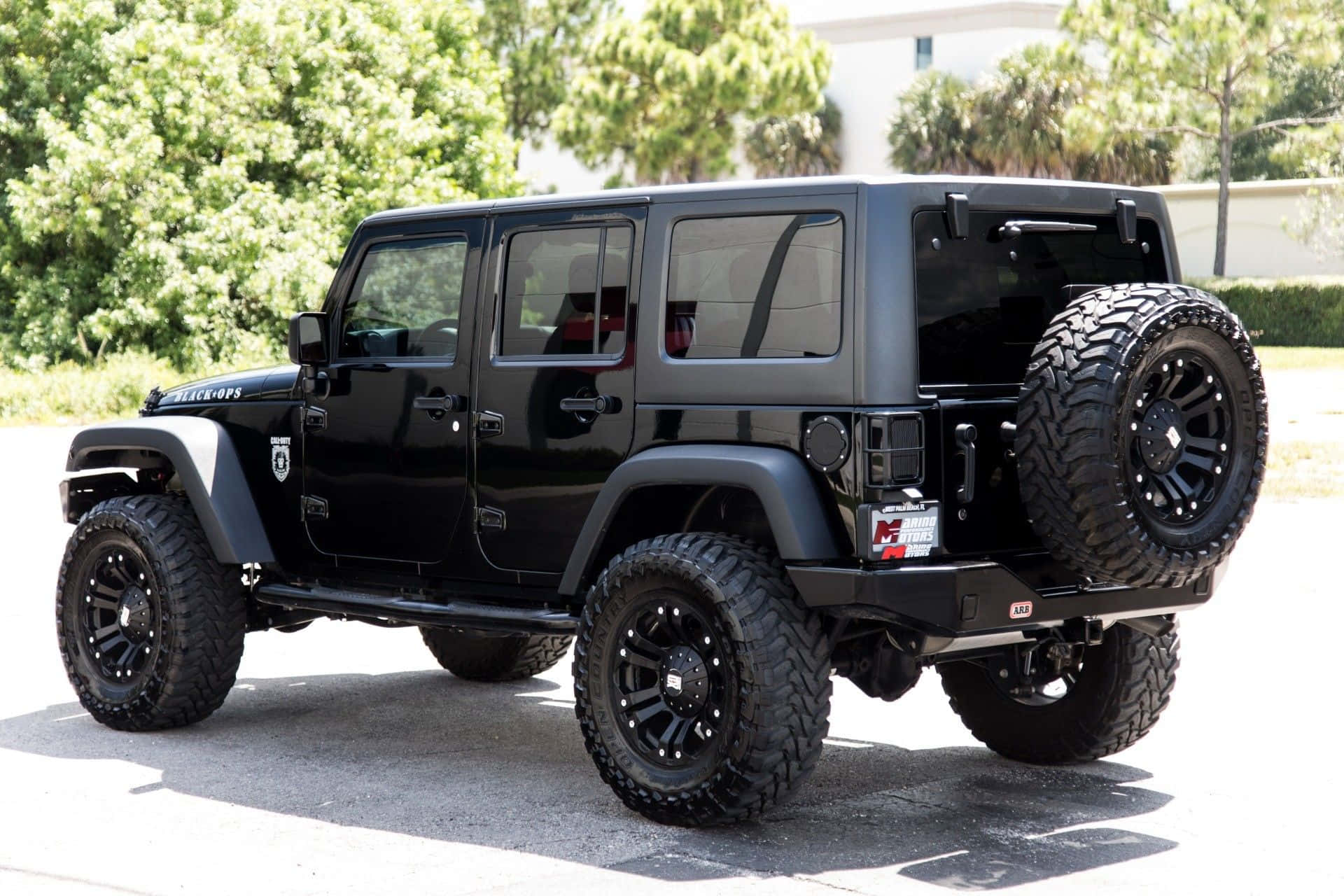 黑色升高底盤的Jeep Wrangler，配備大輪胎，停在陽光普照的瀝青路上，附近有綠樹。