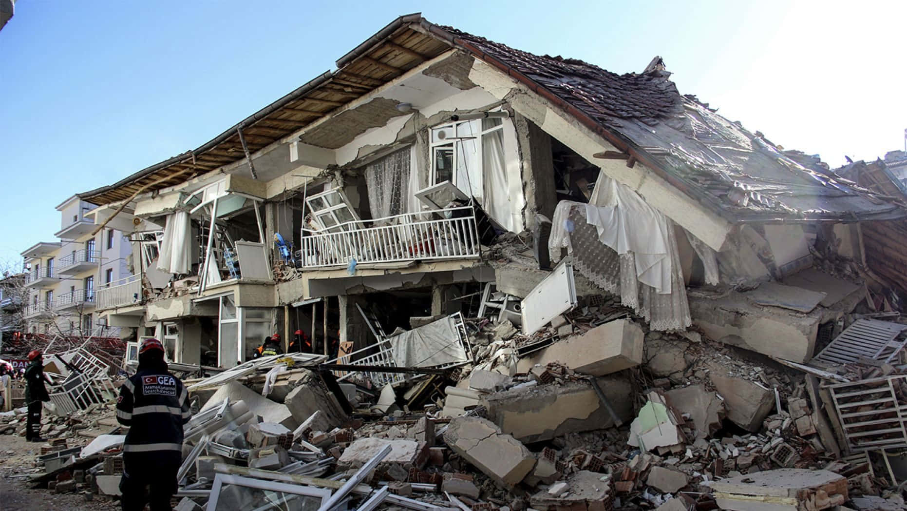 強烈地震撼動東京 壁紙 — 晴朗藍天下，倒塌建築物散落破碎混凝土與瓦礫，救援人員正在評估損毀情況。