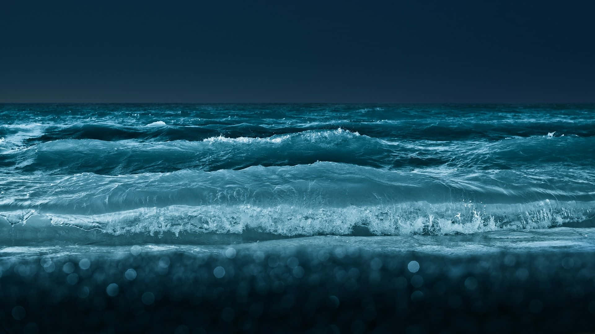 海浪 桌布 壁紙 — 強烈的藍綠色波浪在陰沉的夜空下拍打著黑暗的海岸，前景帶有散景效果。