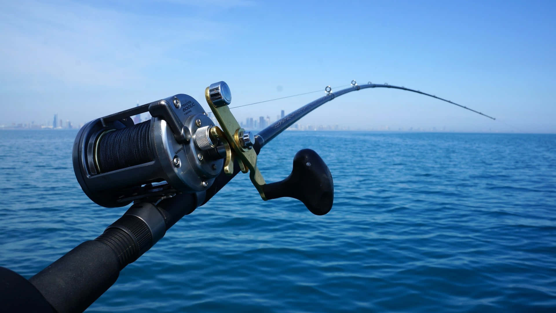 Fishing Rod 桌布 壁紙 — 彎曲的魚竿連捲線器在平靜的藍色水面上拋投，朝向晴朗天空下遠方的城市天際線。