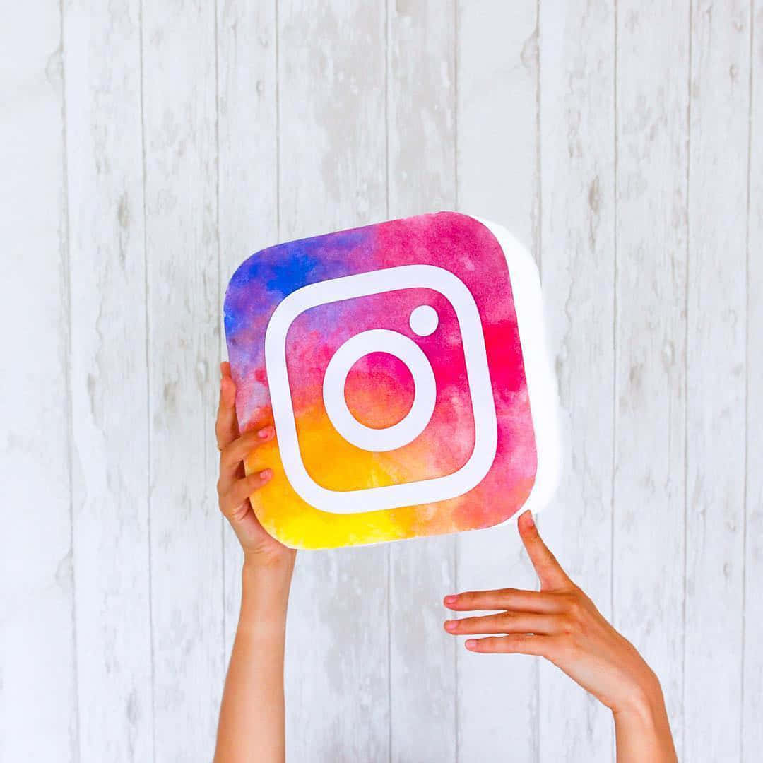 雙手拿著水彩Instagram標誌毛絨玩具，背景是白色木頭。