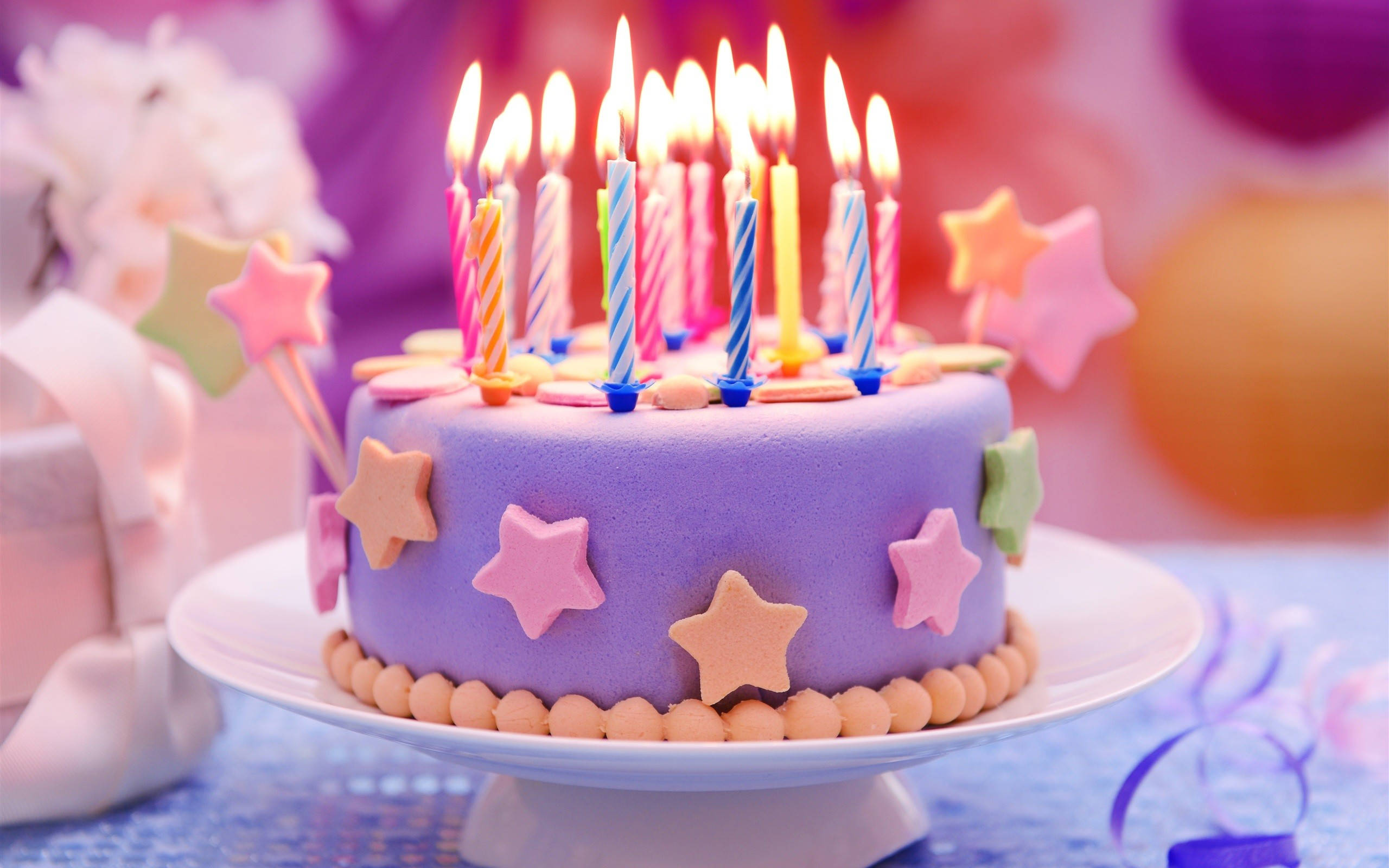 彩色星星生日蛋糕蠟燭 壁紙 — 一個充滿活力的紫色生日蛋糕，裝飾著點燃的蠟燭和色彩繽紛的星星裝飾，放在一個基座上，背景是節日模糊的。