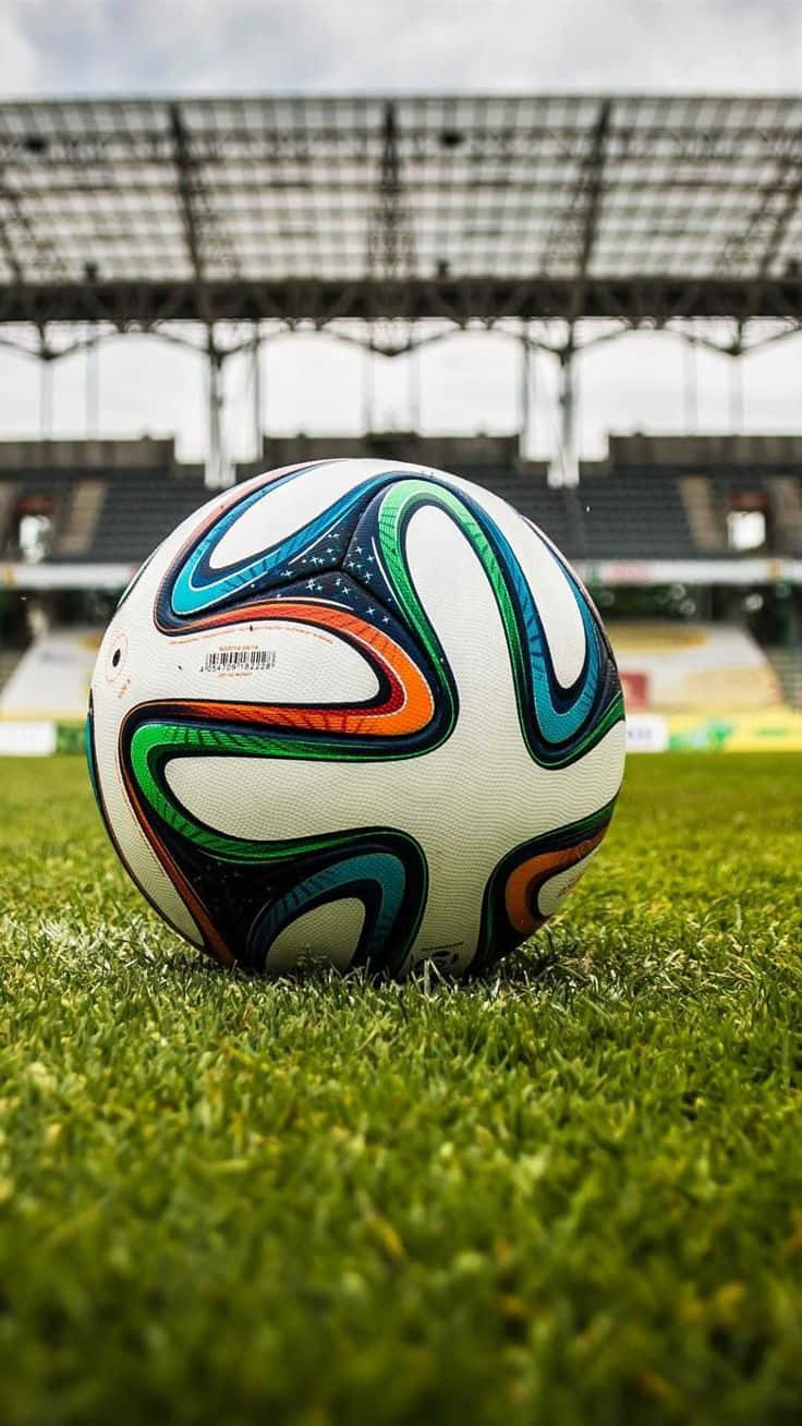 彩色曲線足球背景 壁紙 — 一個色彩鮮豔的 Brazuca 足球放在空蕩蕩的體育館內茂密的綠草上，天空多雲。
