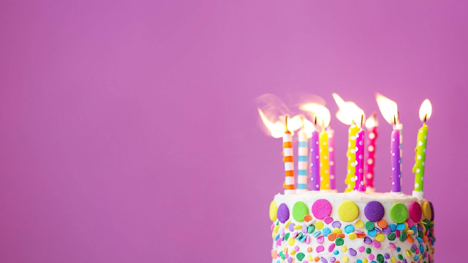 彩色糖果撒粉生日蛋糕 壁紙 — 一個色彩繽紛的生日蛋糕，上面有燃燒的蠟燭和彩虹糖針，背景為純紫色。