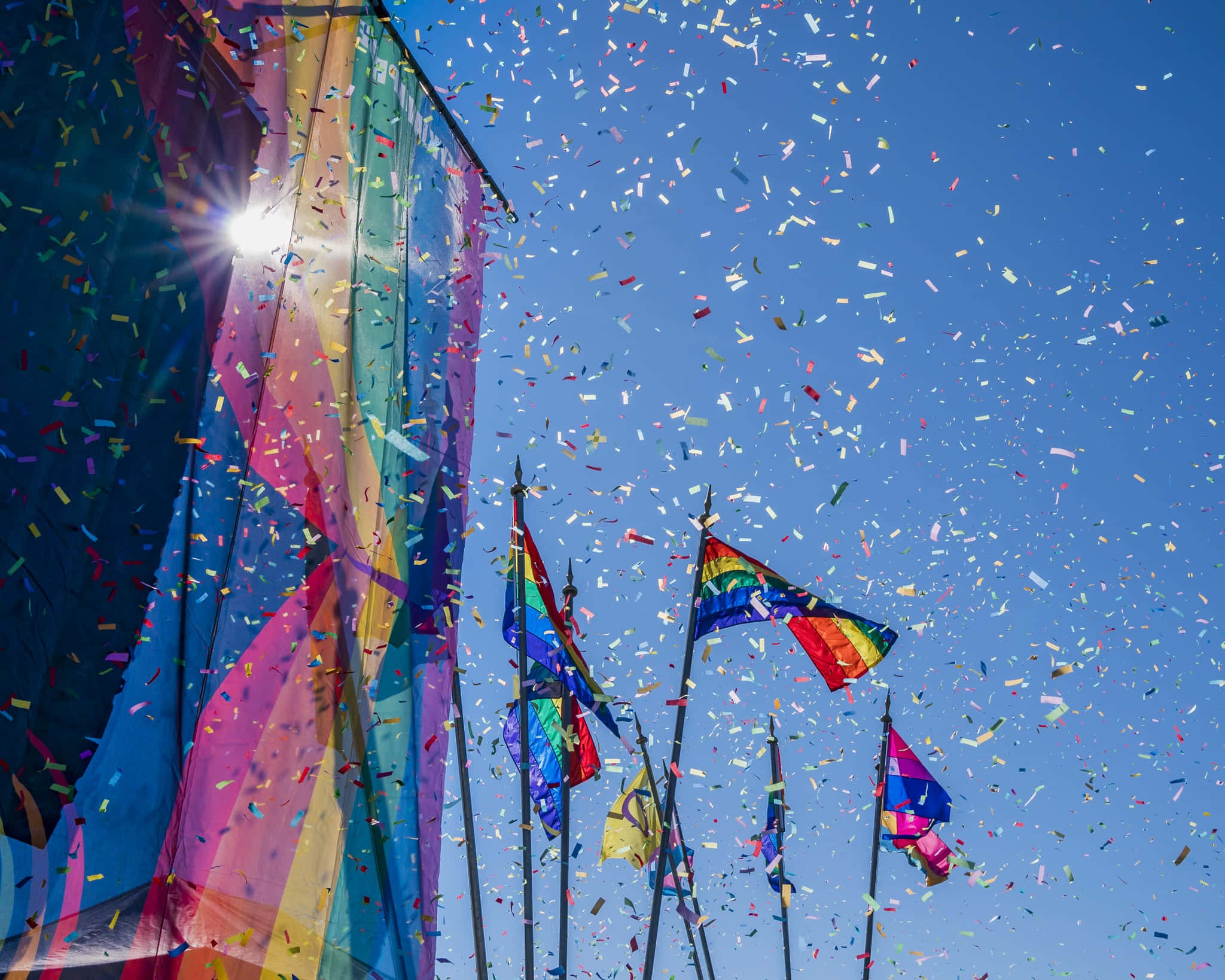 Pride Flag 桌布 壁紙 — 彩虹旗在清澈的藍天下自豪地飄揚，五彩紙屑如雨般灑落慶祝。