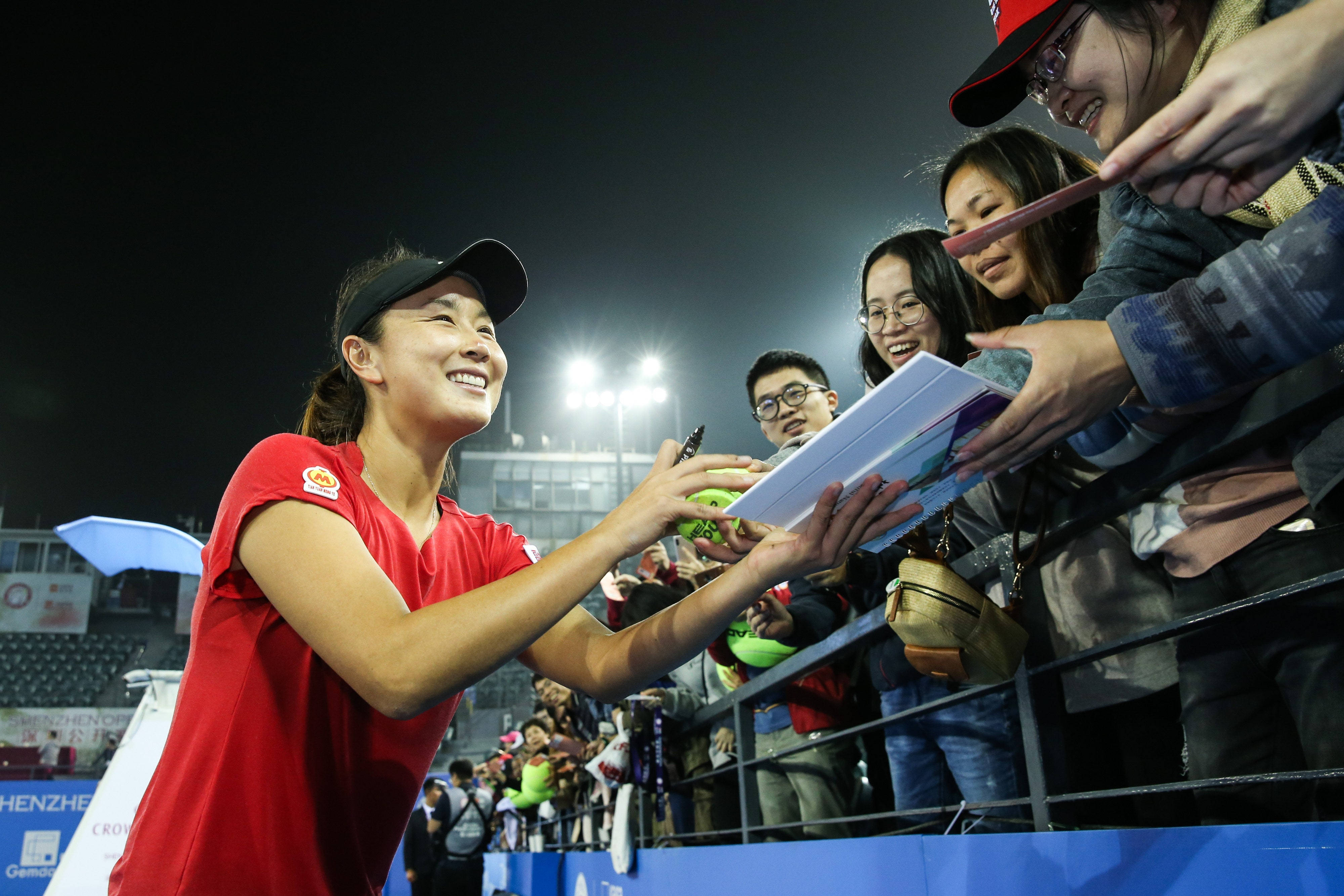 彭帥與粉絲互動 壁紙 — 一位身穿紅色球衣、面帶微笑的女網球選手在夜間明亮的體育場燈光下為球迷簽名。