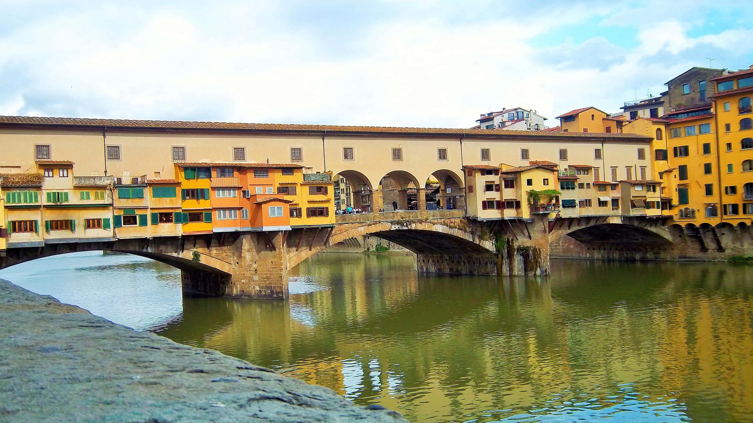 彭特維喬橋附近酒店 壁紙 — 佛羅倫斯的舊橋（Ponte Vecchio）拱跨阿諾河，石造結構兩旁排列著色彩繽紛的商店，天空部分多雲。