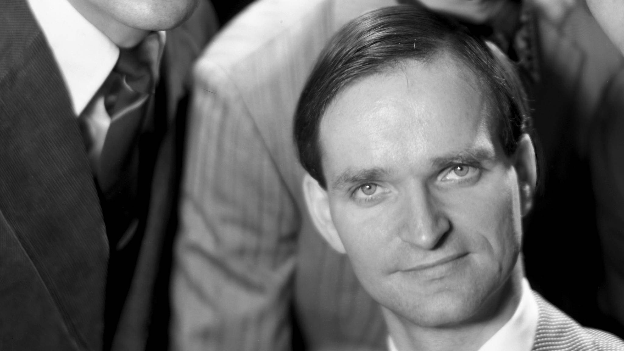 影響深遠的先驅：Kraftwerk 成員 Florian Schneider 壁紙 — 一個穿著西裝的男人黑白特寫肖像，帶著微妙微笑直視鏡頭。