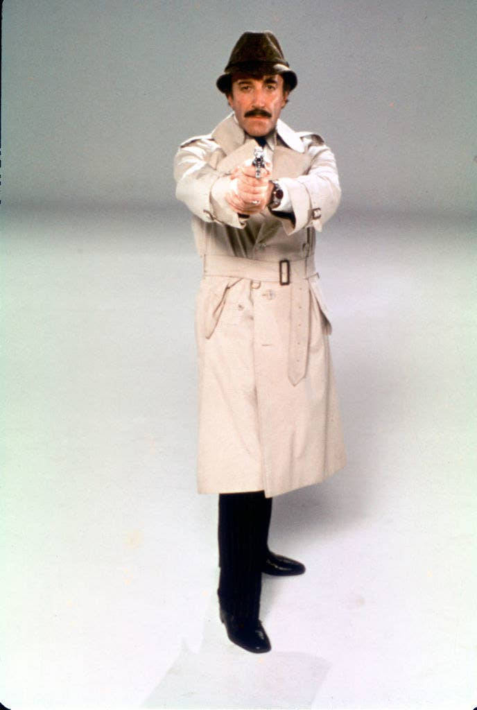 彼得·塞勒斯飾演 Inspector Clouseau 壁紙 — 一名身穿米色風衣和軟呢帽的男子，在純色攝影棚背景下，直接將手槍瞄準觀眾。