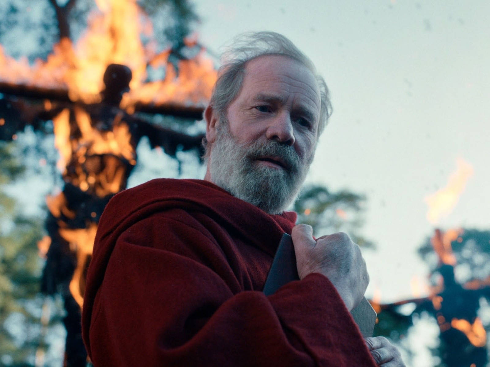 彼得·馬蘭在Netflix《詛咒》中飾演神父卡登 壁紙 — 一個鬍鬚男人身穿紅色長袍，在黃昏的森林中站在一個燃燒的木製肖像前，手持一把刀。