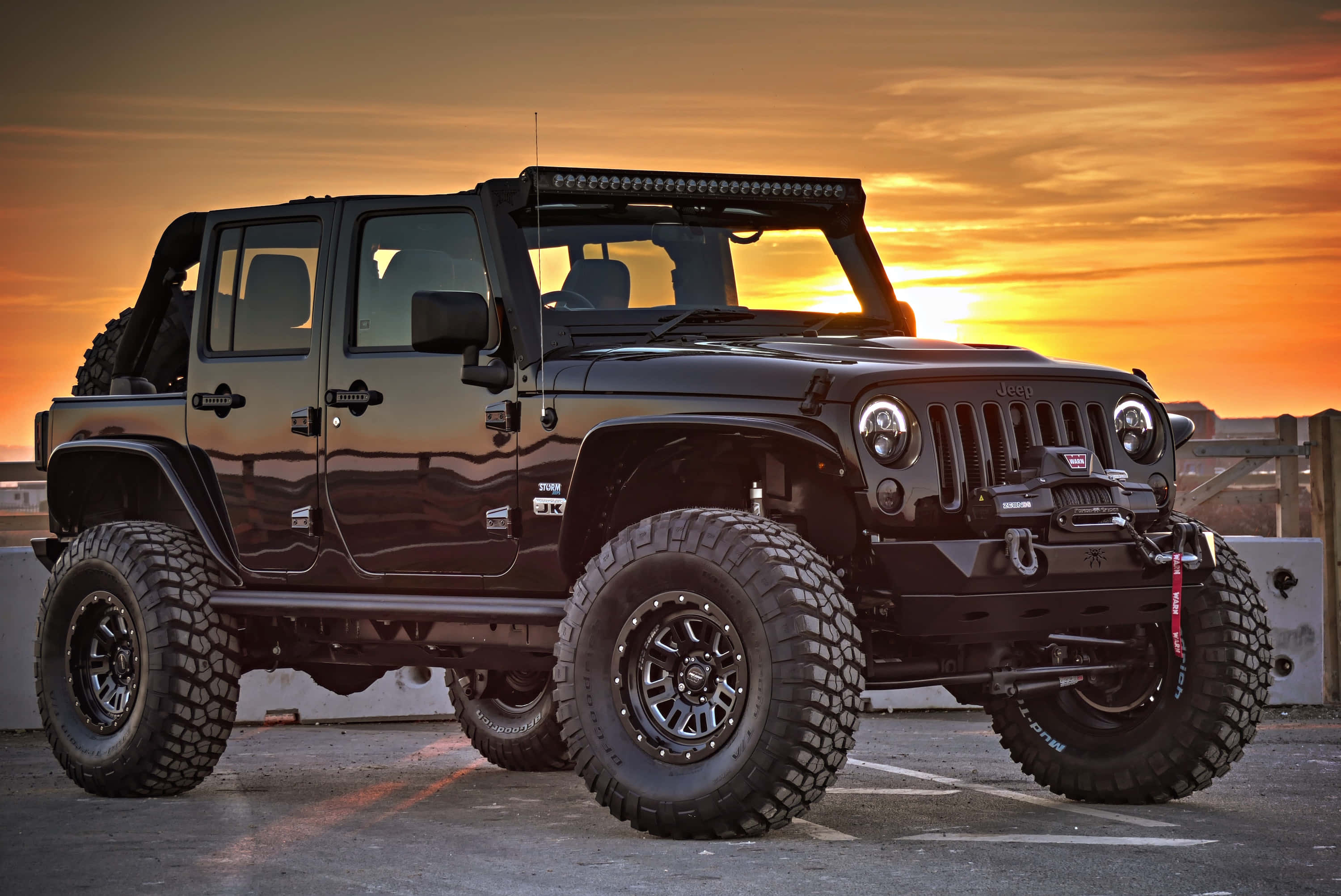 征服任何地形，體驗Jeep Wrangler的非凡魅力 壁紙 — 一輛黑色升高嘅Jeep Wrangler，配超大輪胎，喺充滿活力嘅橙色日落時分停泊喺停車場。