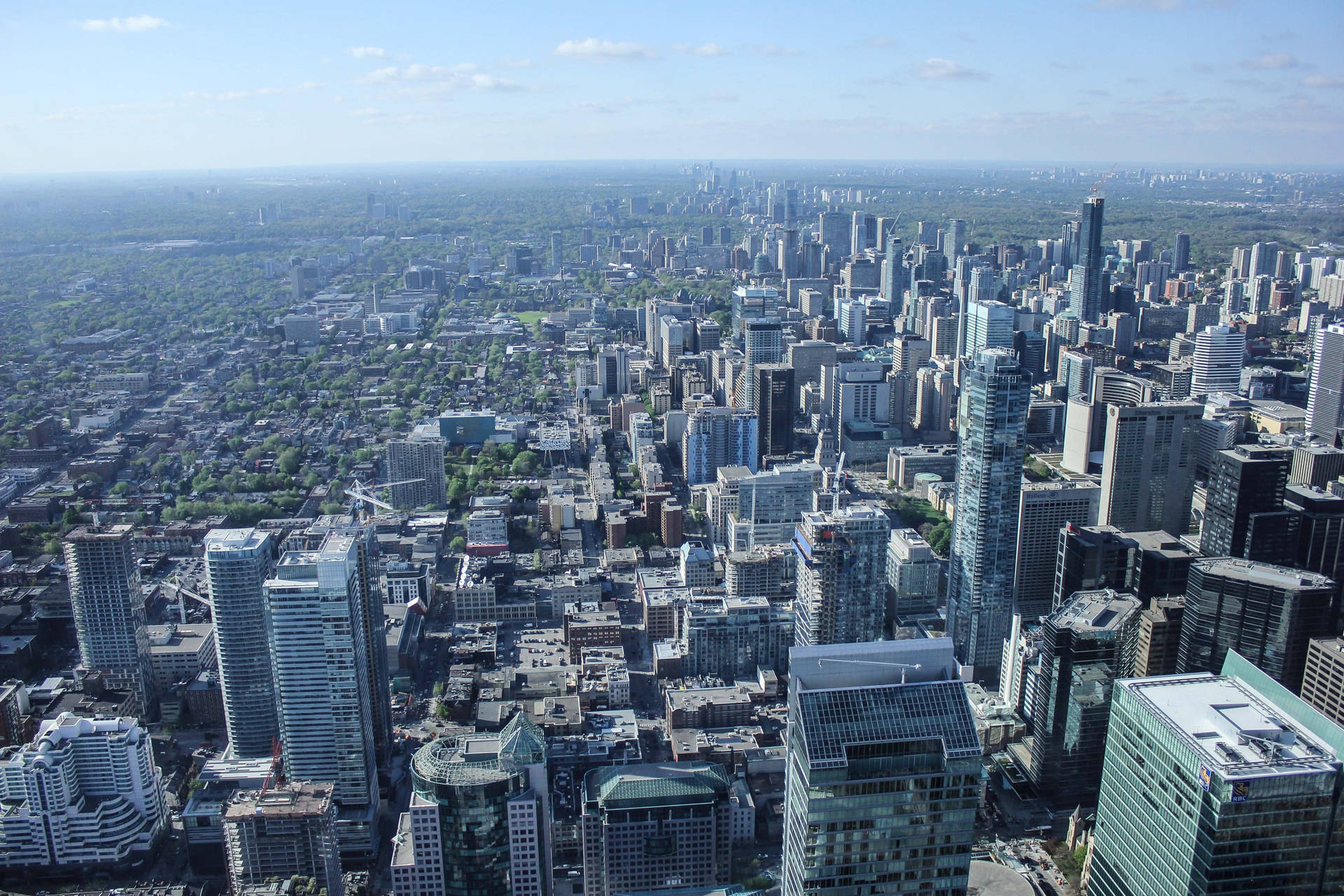 從CN塔觀景台俯瞰多倫多市景 壁紙 — 從高空俯瞰，在明亮的藍天下，一座現代化城市景觀延伸開去，玻璃摩天大樓與綠色公園交織。