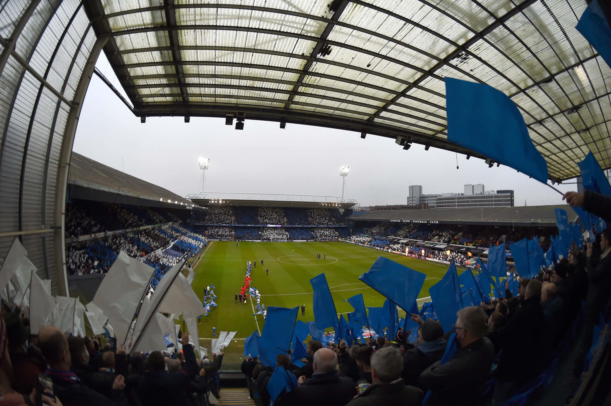 Ipswich Town 桌布 壁紙 — 從一個擁擠的體育場的魚眼視角，球迷在陰天下的足球比賽中揮舞著藍色和白色的旗幟。