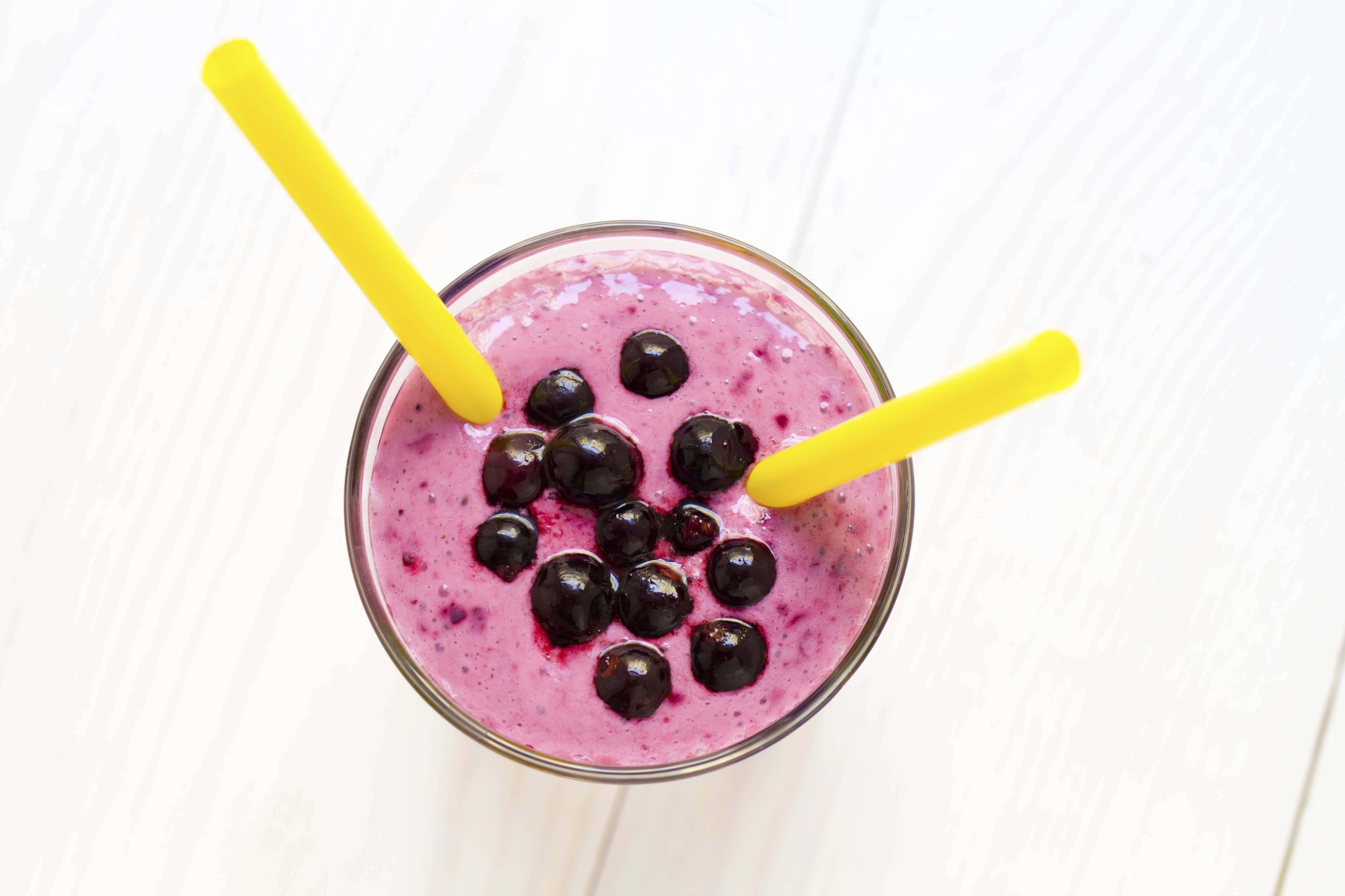 Blueberry Smoothie 桌布 壁紙 — 從上方俯瞰，一個充滿活力的紫色藍莓奶昔，上面放著新鮮藍莓和兩根黃色吸管，置於白色木質表面上。
