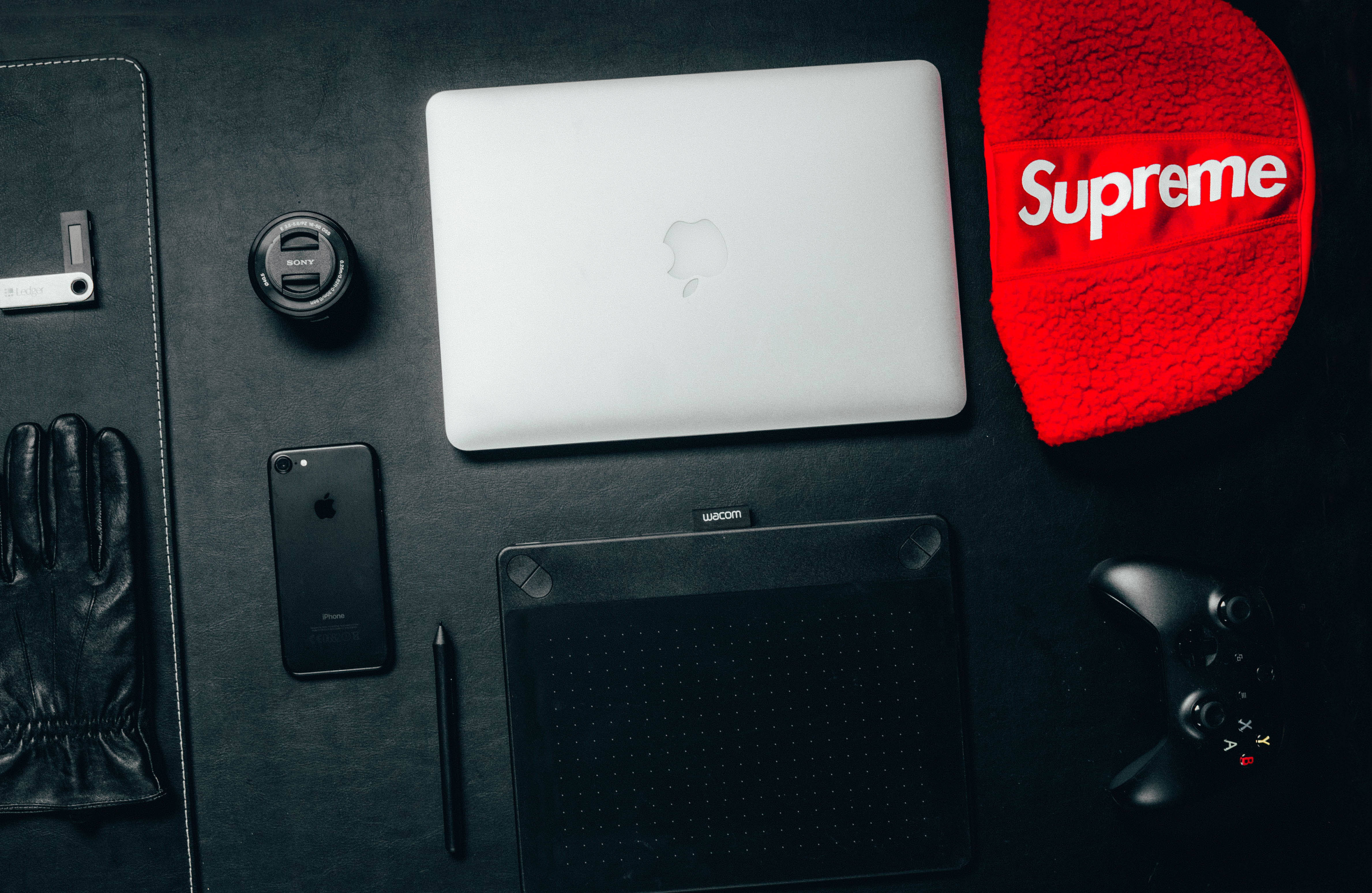 supreme 桌布 壁紙 — 從上方俯瞰一個時尚的科技設置，包括銀色筆記型電腦、黑色手機、繪圖板、遊戲控制器和紅色Supreme毛帽，放置在深色表面上。