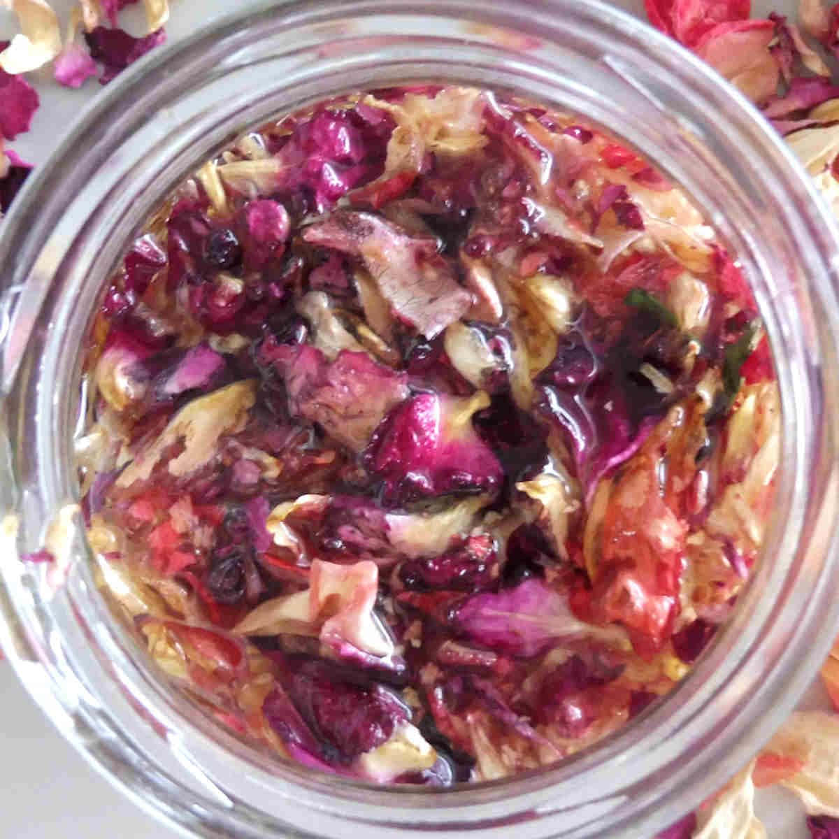 Rose Oil 桌布 壁紙 — 從上方俯瞰一個玻璃罐，裡面裝滿了深紫色和紅色的玫瑰花瓣，在琥珀色液體中浸泡，周圍散落著乾燥的花瓣。