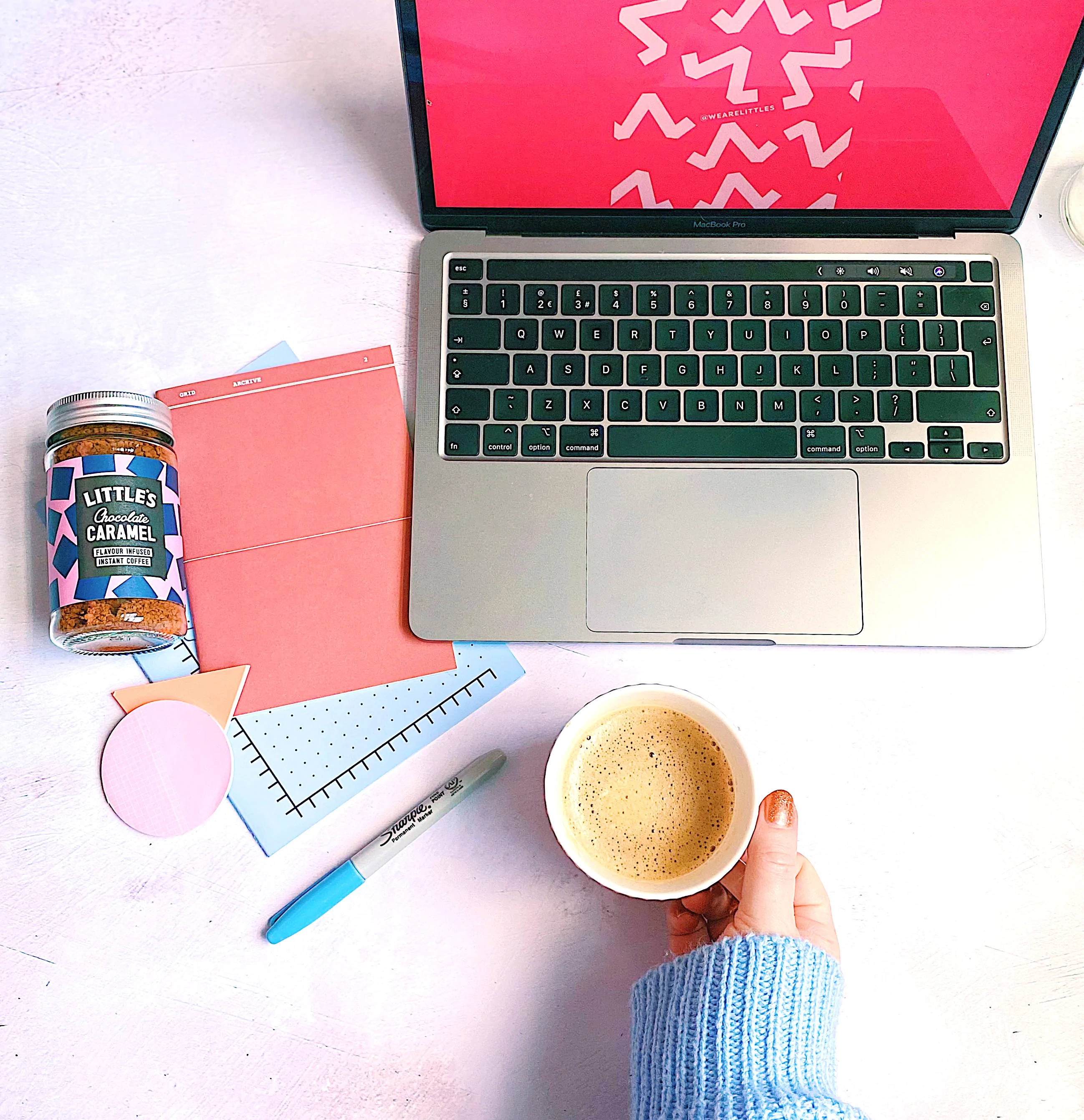 Coffee Aesthetic 桌布 壁紙 — 從上方俯瞰一隻手在白色桌子上拿著咖啡，旁邊有手提電腦、焦糖罐和彩色文具。