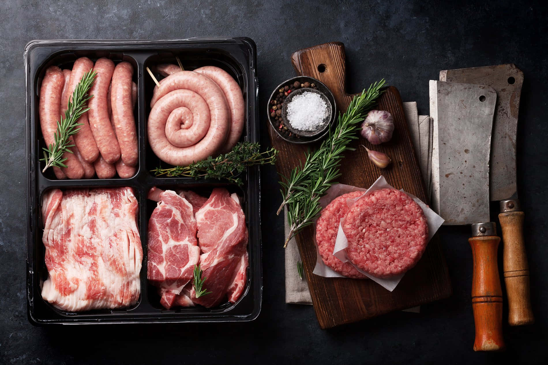 meat 桌布 壁紙 — 從上方俯瞰的視角，展示生肉，包括香腸、肉餅和煙肉，與香草、鹽、大蒜和切肉刀一同排列在深色紋理表面上。