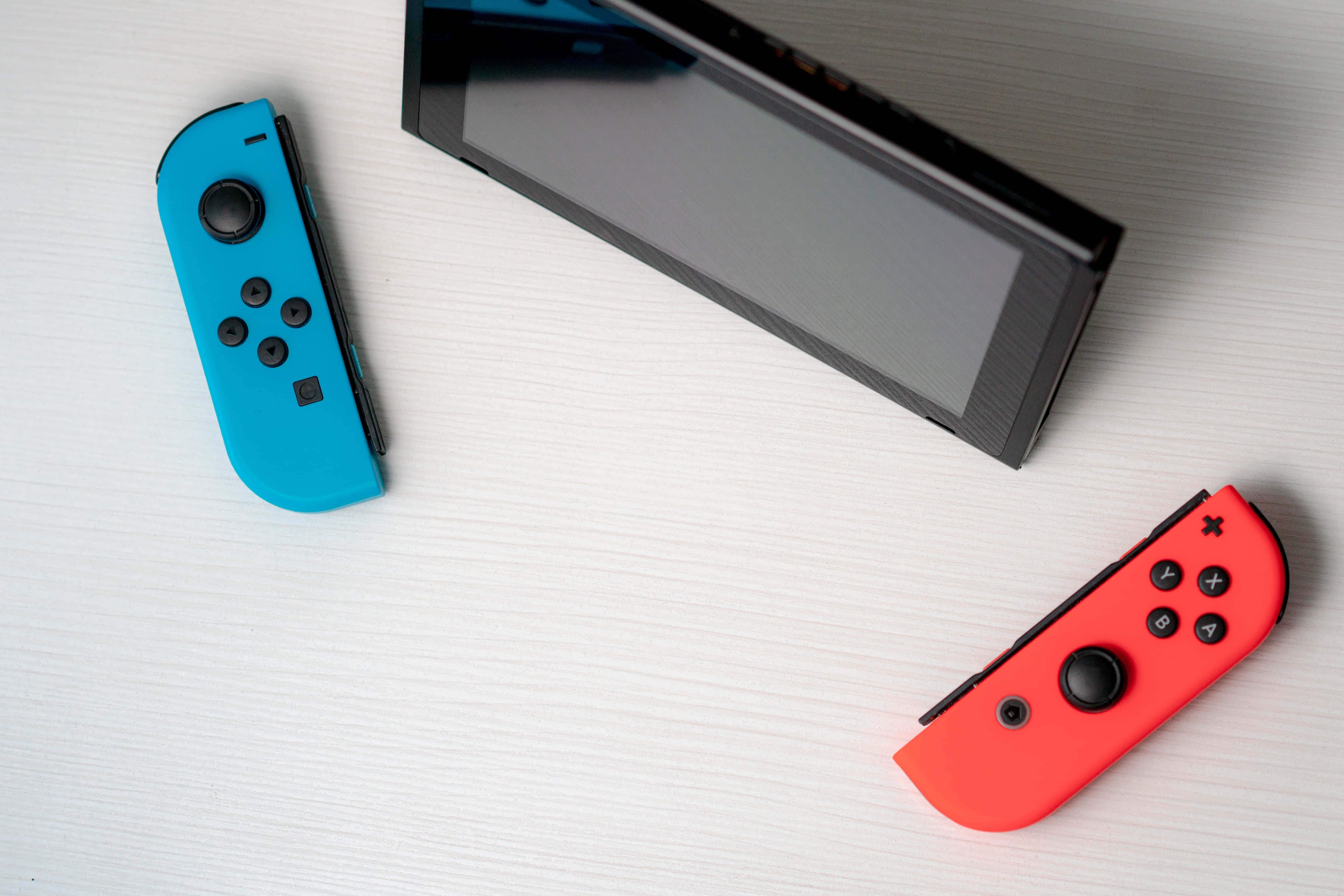 Gaming Consoles 桌布 壁紙 — 從上方俯瞰黑色Nintendo Switch主機，配備藍色和紅色Joy-Con控制器，放置在淺色木質表面上。