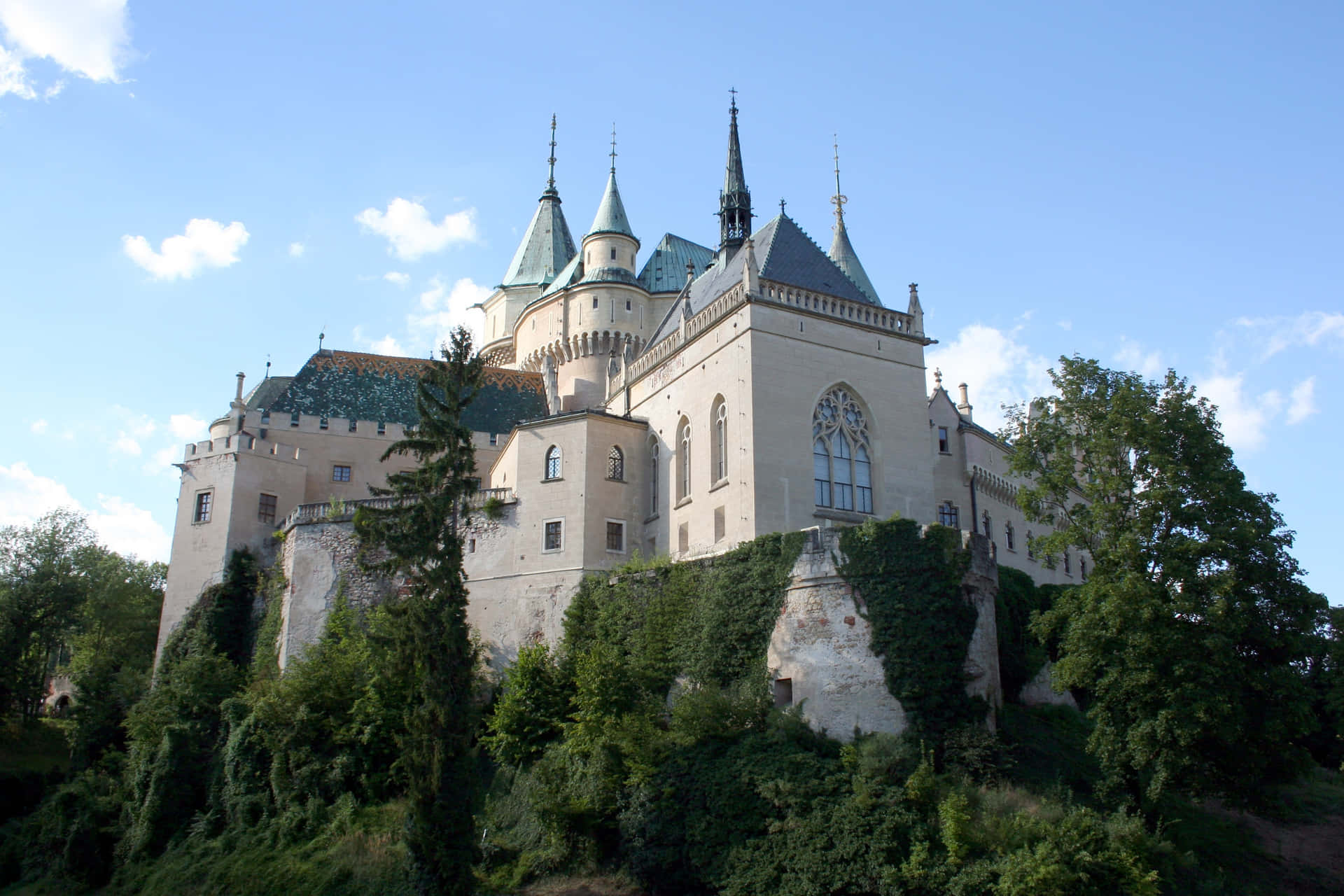 從下方仰望波亞尼奇城堡 壁紙 — 一座宏偉的石砌城堡，擁有綠色瓦片尖頂，雄偉地聳立在茂密的山丘上，上方是點綴著蓬鬆白雲的明亮藍天。