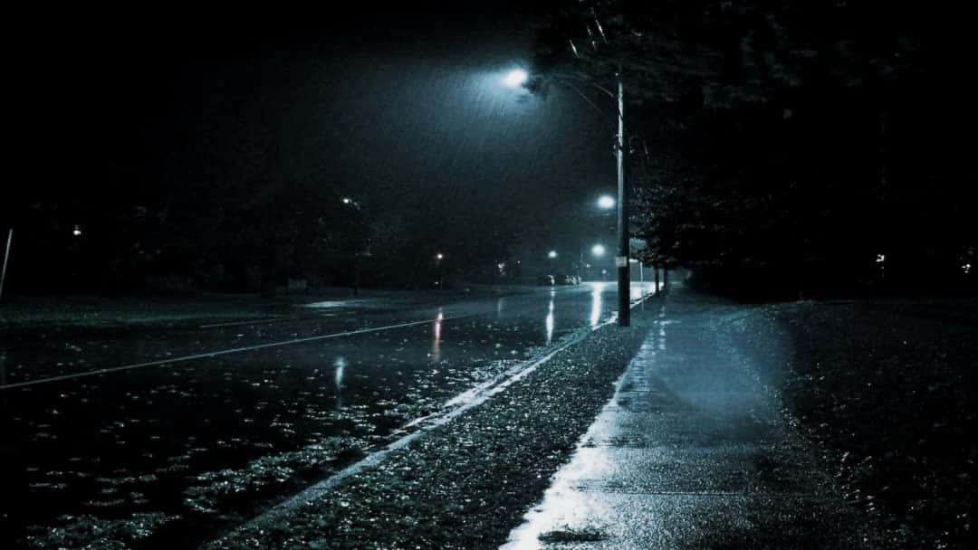 從任何地方欣賞繁星夜景 壁紙 — 一條被雨水打濕的都市街道，夜晚被刺眼的街燈照亮，在落下的雨中投下長長的影子。