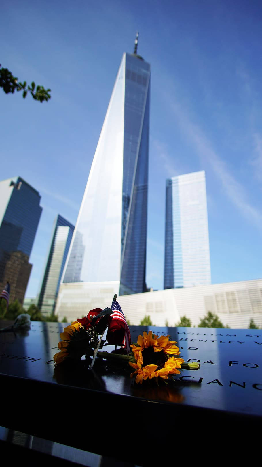 911 Memorial 桌布 壁紙 — 從低角度觀看世界貿易中心一號樓，背景是晴朗的藍天，前景的紀念牌上擺放著向日葵和美國國旗。