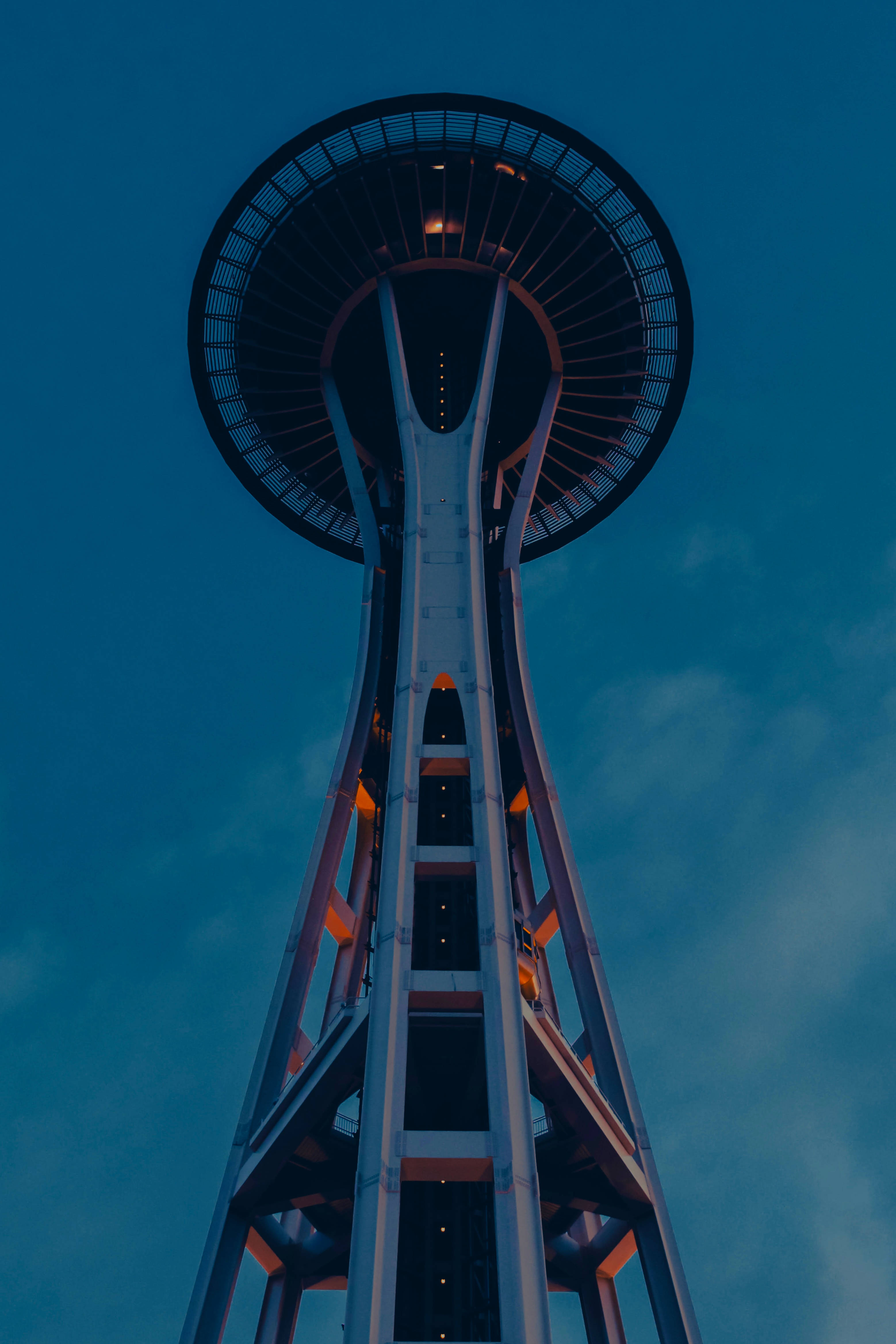 Seattle iPhone Space Needle Structure 壁紙 — 從低角度觀賞太空針塔在黃昏時分，溫暖的燈光在深藍色天空下閃耀。