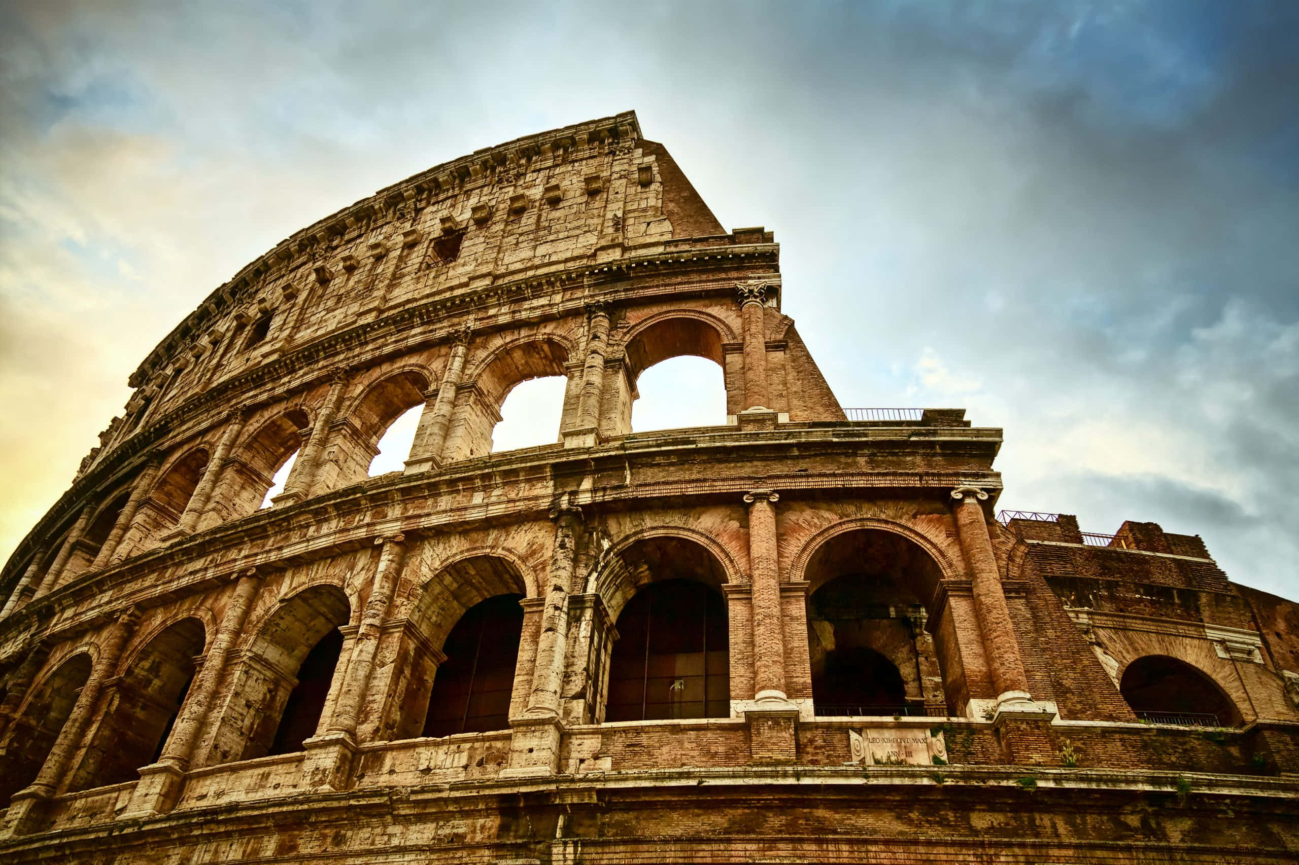 Roman 桌布 壁紙 — 從低角度觀賞羅馬古羅馬競技場，天空多雲，戲劇性十足，溫暖的金色光線。