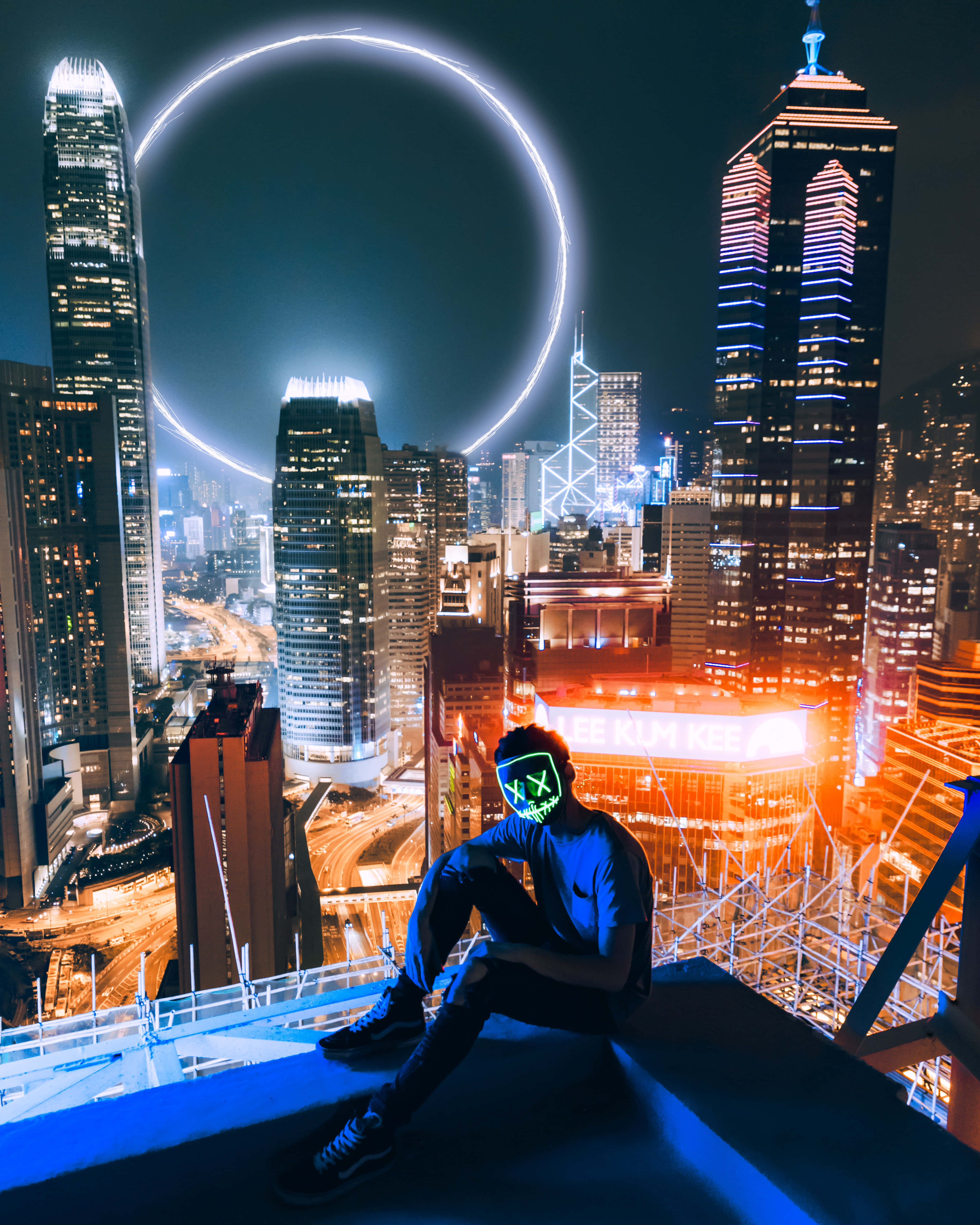 從天台欣賞美麗景色 壁紙 — 一個人戴著發光綠色面具坐在摩天大樓邊緣，俯瞰著霓虹燈閃爍的城市景觀，夜空中有發光的圓形光環。