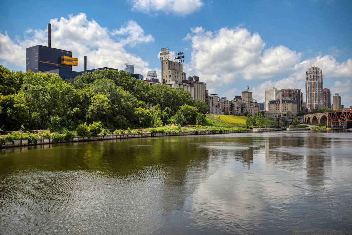 從天際線俯瞰密西西比河 壁紙 — 充滿活力的城市景觀，河流倒映著摩天大樓和茂密的綠樹，天空湛藍，白雲朵朵。