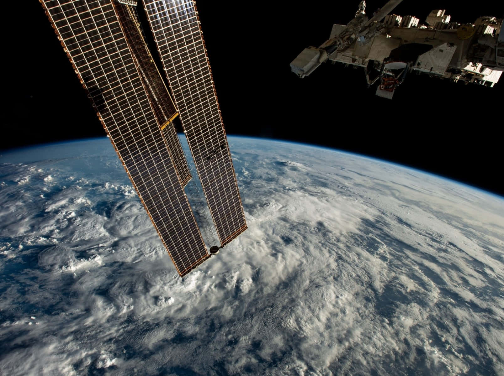 從太空衛星觀看地球 壁紙 — 太陽能電池板和太空站模組在旋轉的藍白地球上方，向著黑色虛空延伸。