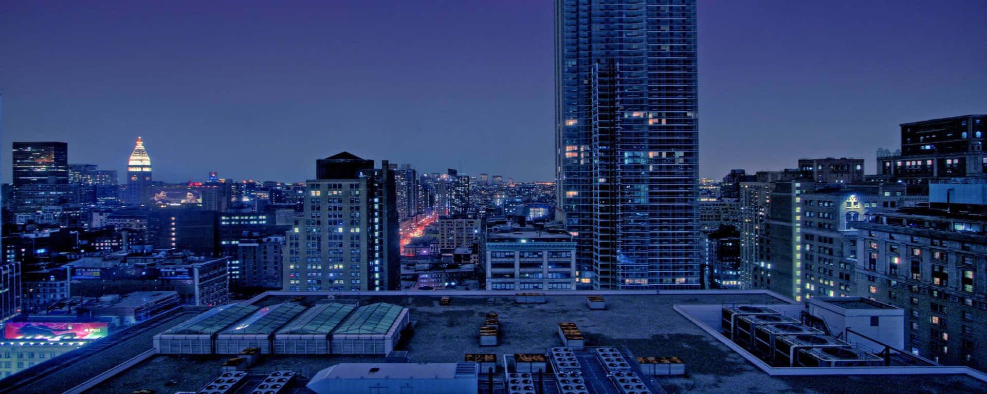 城市景觀 桌布 壁紙 — 從屋頂俯瞰黃昏時分的城市天際線，窗戶閃爍著光芒，遠處的塔樓在深紫色天空下被照亮。