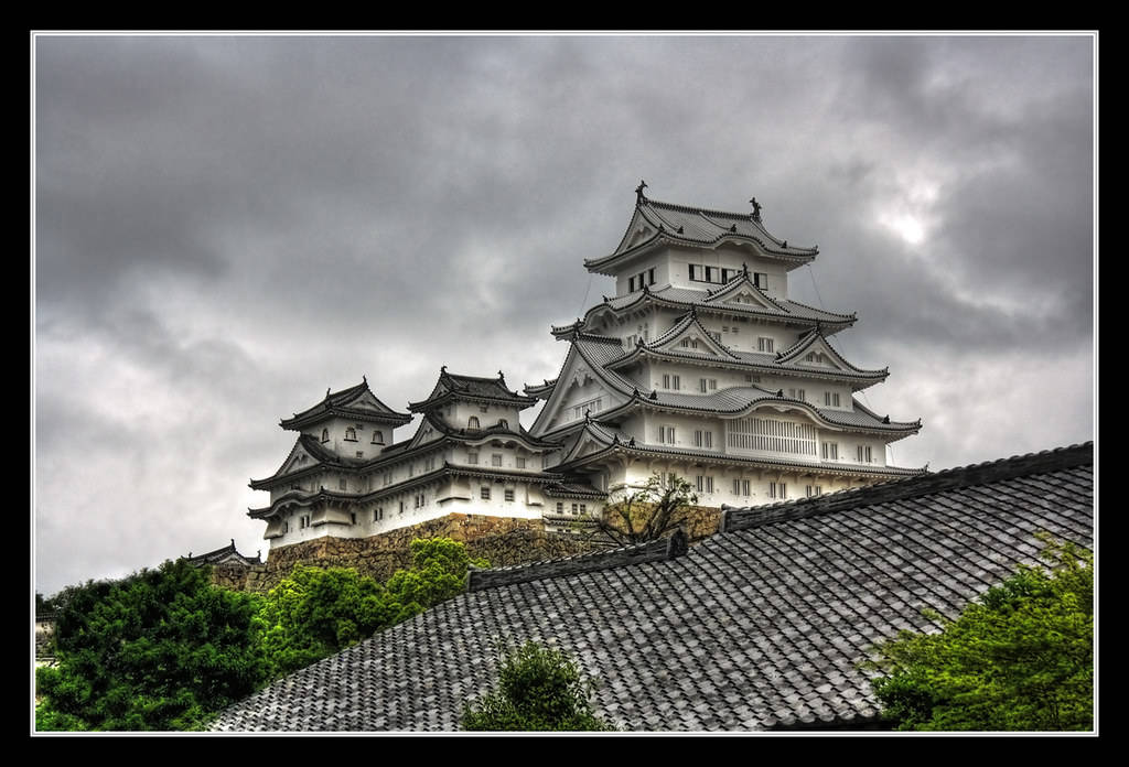 山頂上的白色日本城堡，深色瓦片屋頂和綠樹，多雲天空。