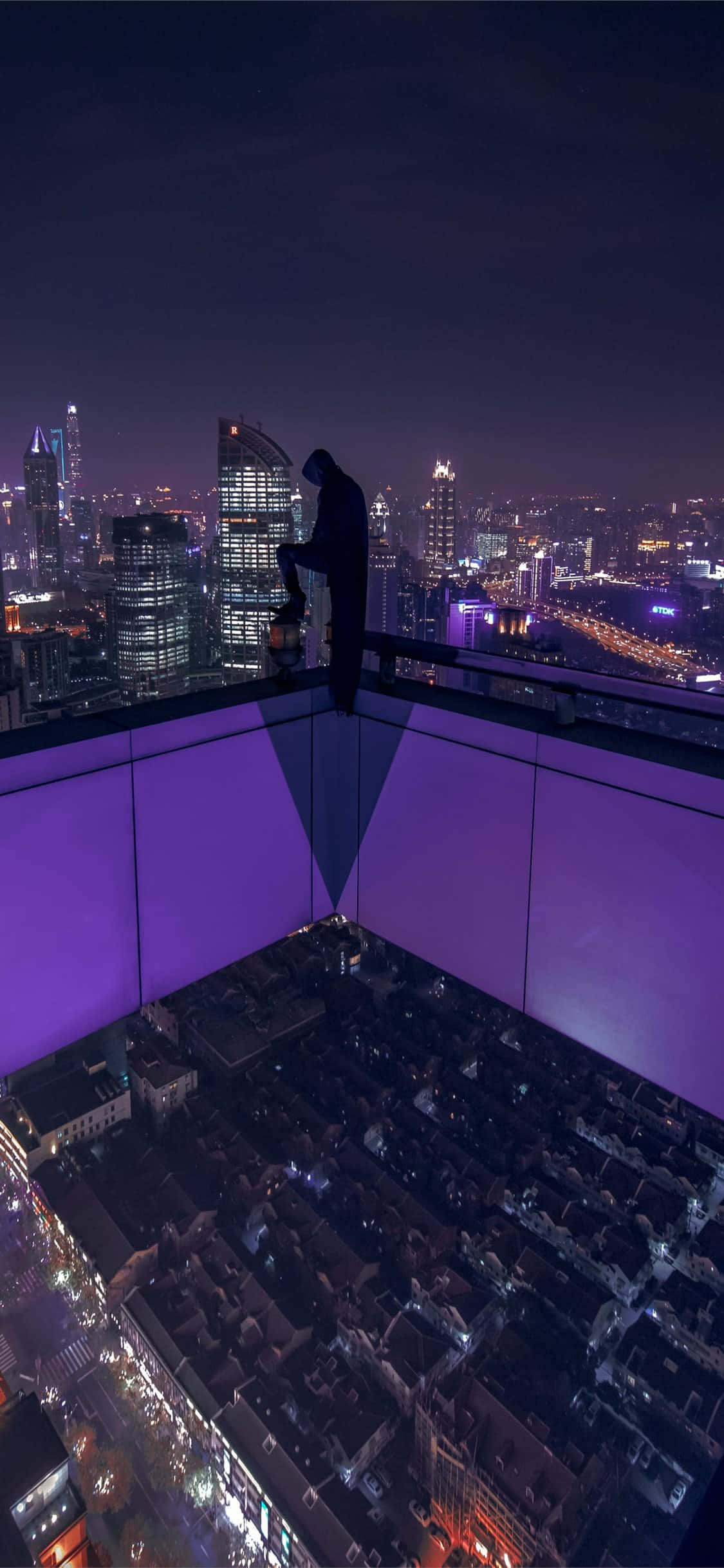 從屋頂欣賞城市天際線夜景 壁紙 — 一個孤獨的身影站在屋頂上，俯瞰著夜晚充滿活力的霓虹燈城市景觀，帶有紫色環境燈光。