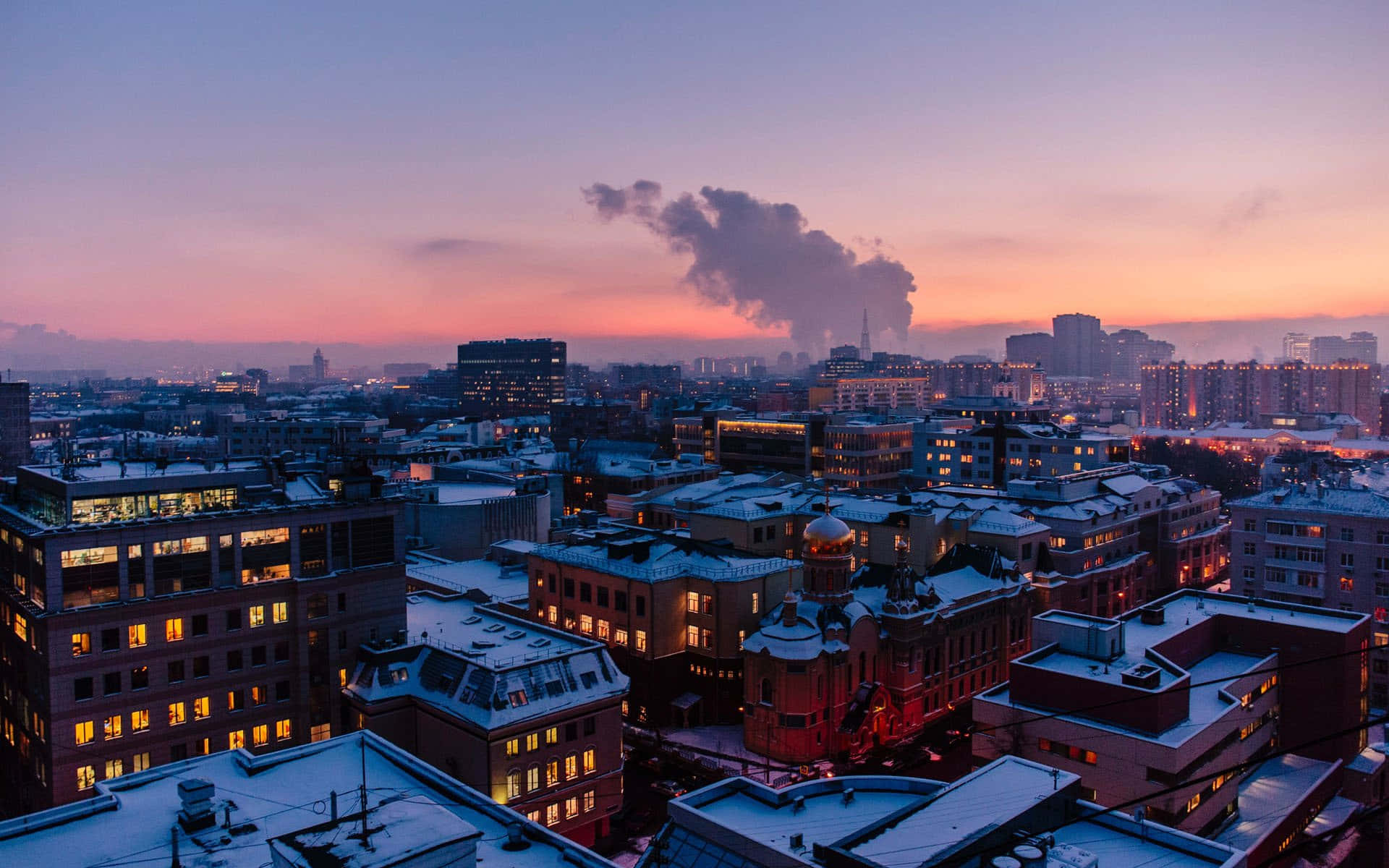 從屋頂欣賞壯麗城市風景 壁紙 — 白雪覆蓋屋頂，城市燈光在帶有粉紅和紫色色調的暮色天空中閃耀。