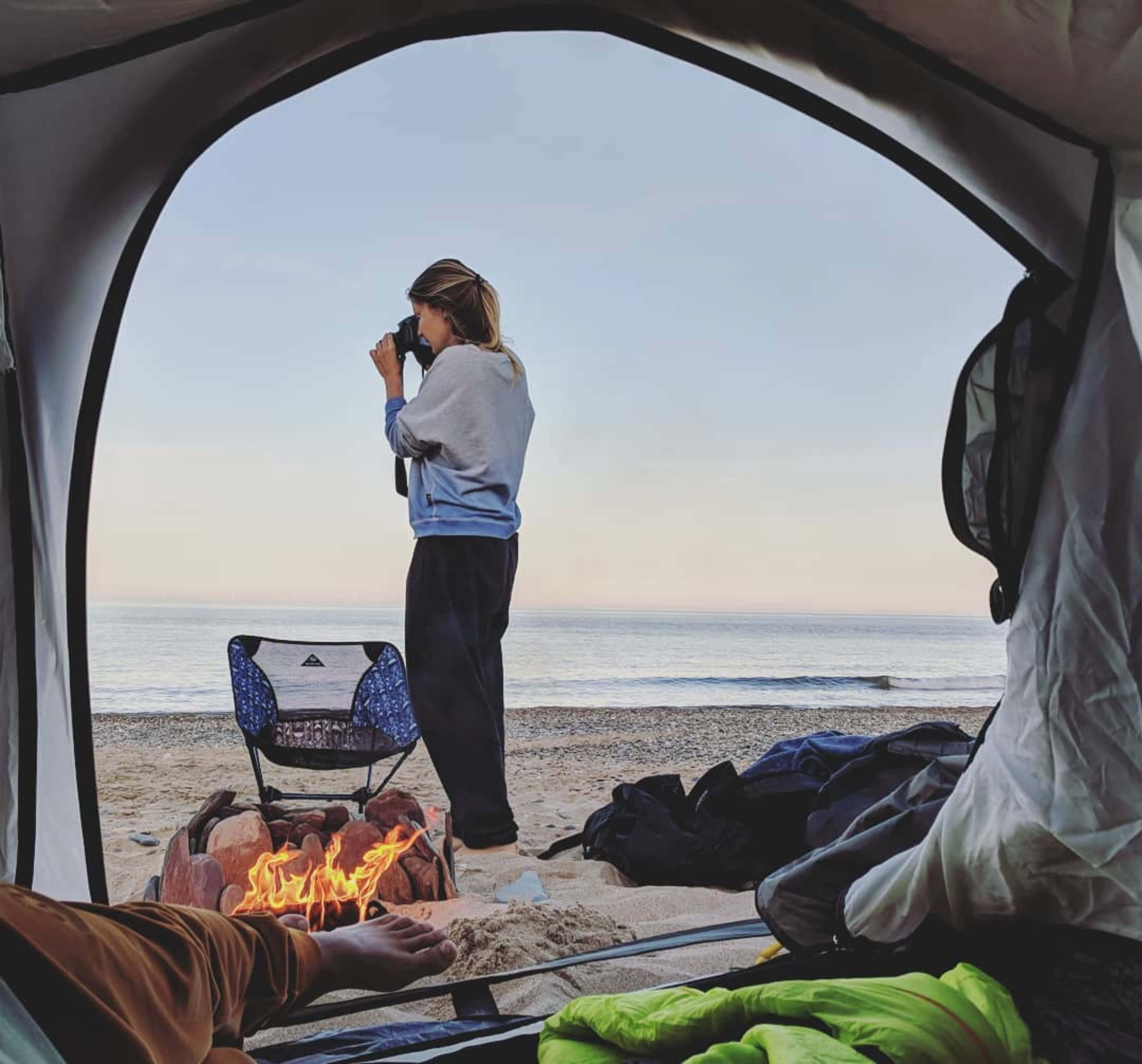 Minimalist Camping 桌布 壁紙 — 從帳篷內部，一個人站在海灘營火旁拿著相機，而另一個人則在附近休息雙腳。