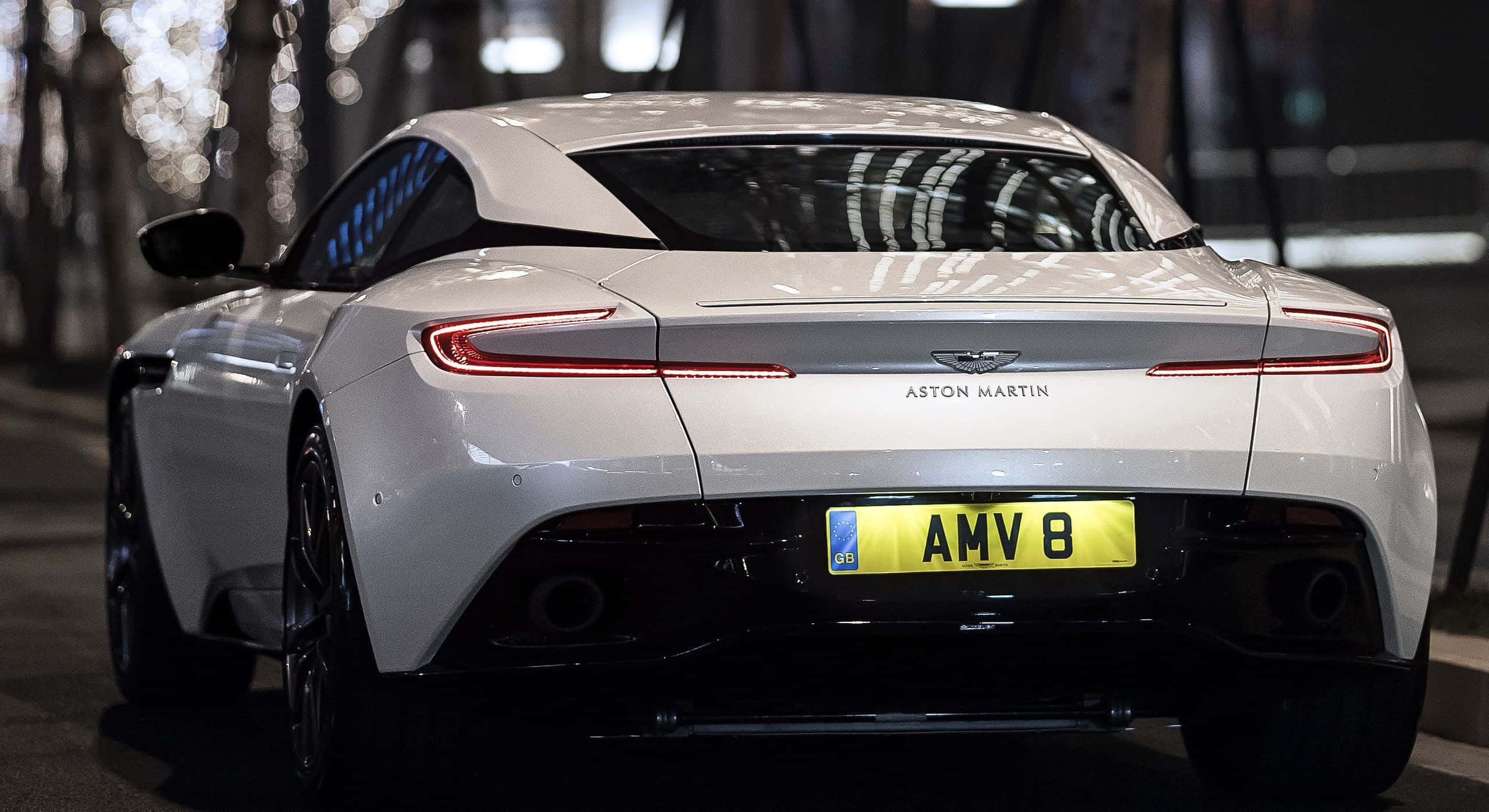 Aston Martin Vanquish 桌布 壁紙 — 從後方觀看一輛時尚的白色Aston Martin跑車，夜間帶有發光的尾燈和模糊的城市燈光背景。