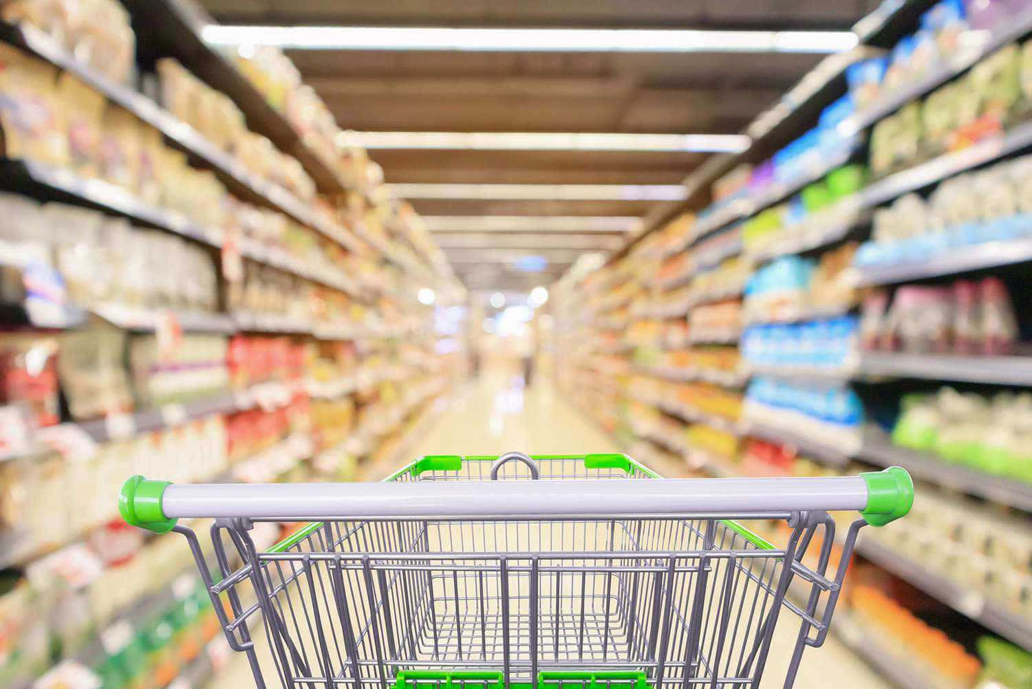 Grocery Store 桌布 壁紙 — 從正面看的購物車在明亮燈光的超市通道中行駛，通道兩旁擺滿色彩繽紛的產品貨架。