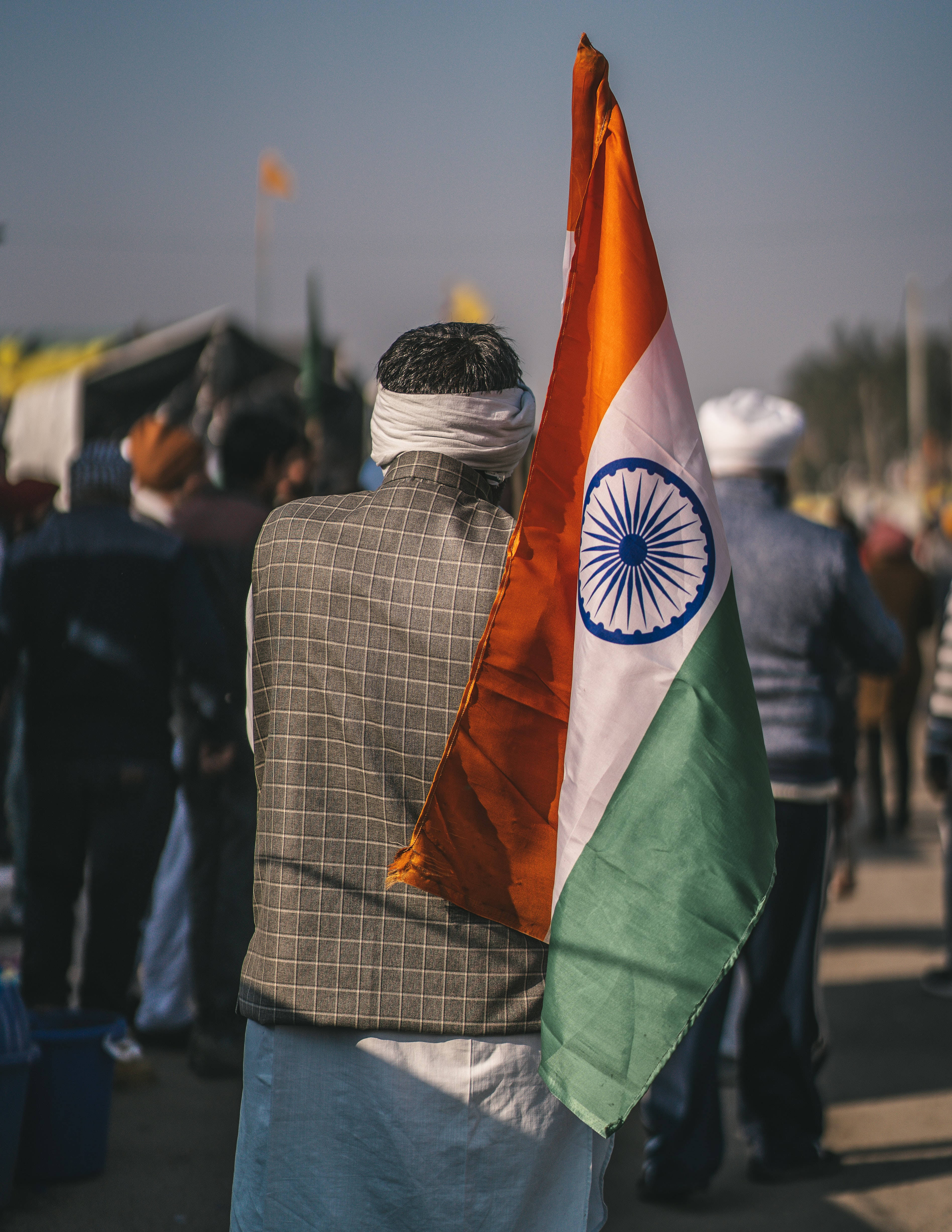 Indian 桌布 壁紙 — 從背後看到一名男子在擁擠的戶外聚會中，在明亮的日光下將一面大型印度國旗扛在肩上。
