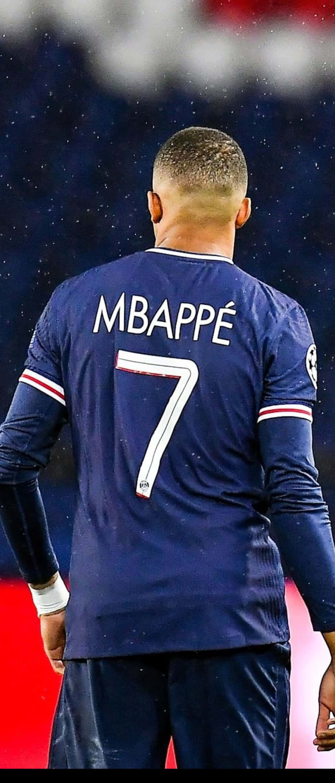 Mbappe 桌布 壁紙 — 從背後看身穿藍色7號球衣的足球員麥巴比站在球場燈光下的雨地球場上。