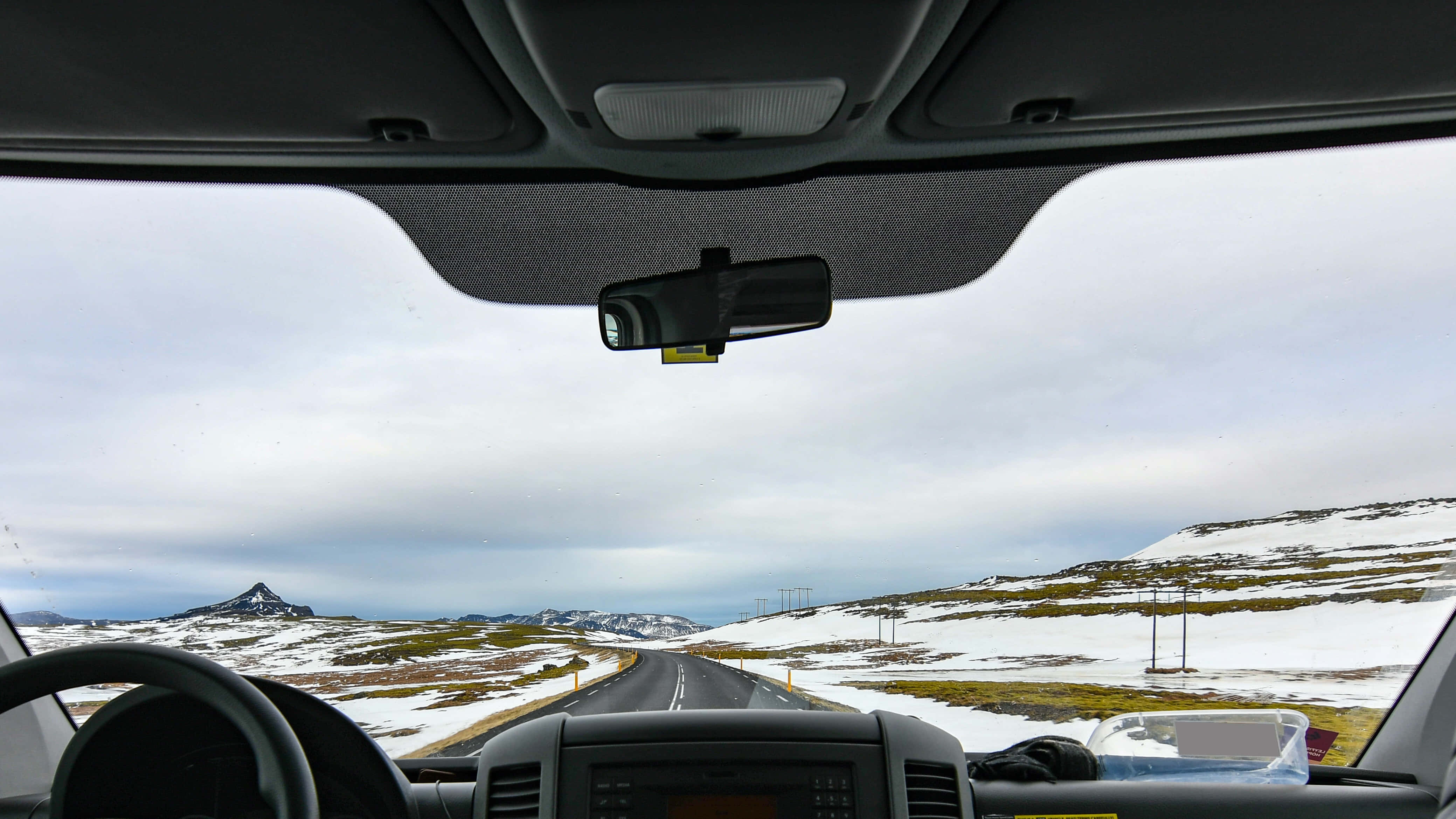 road trip 桌布 壁紙 — 從車內沿著積雪的山路行駛，遠處山峰在陰天之下。