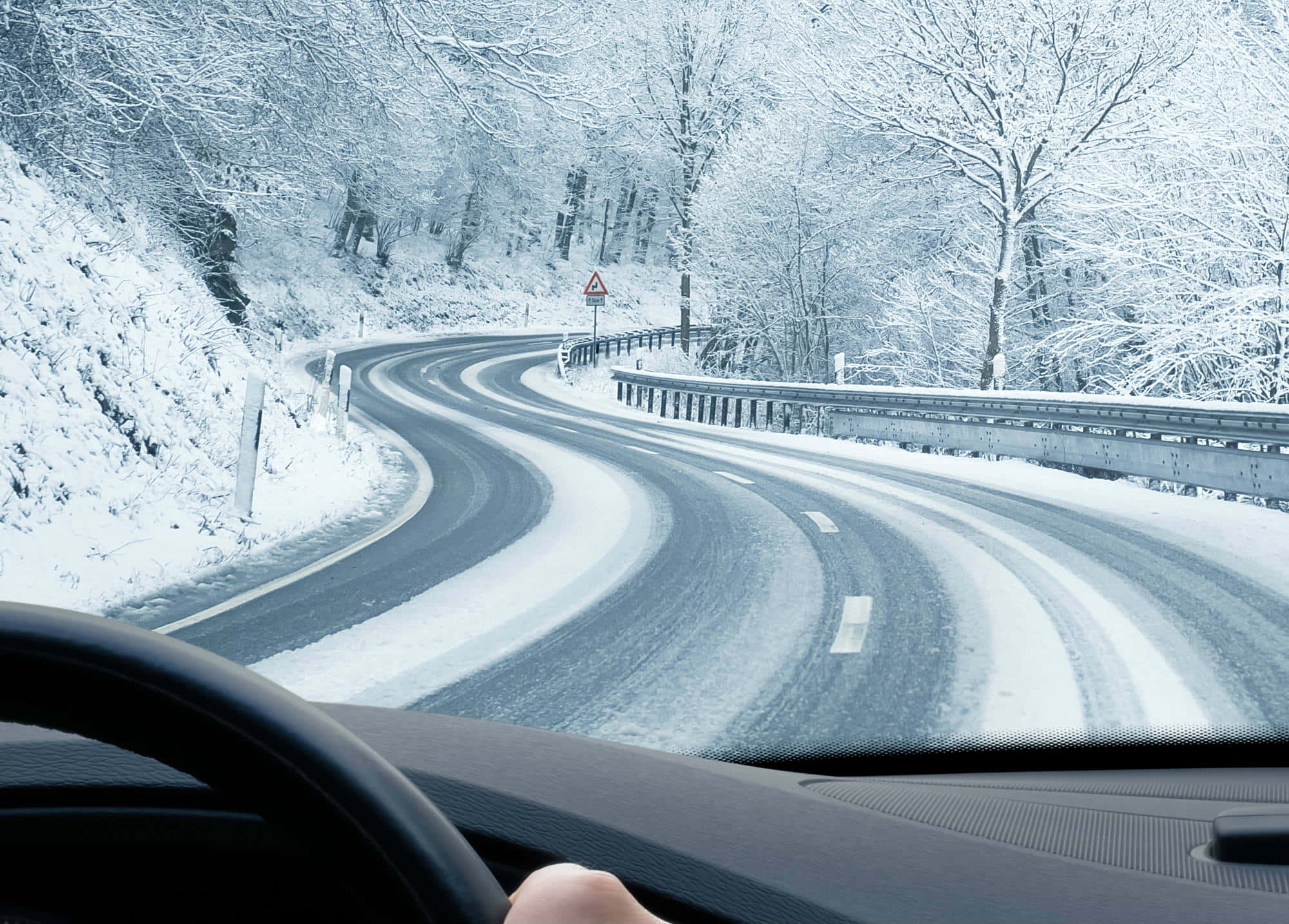 Winter Driving 桌布 壁紙 — 從車內看到一隻手放在方向盤上，一條蜿蜒的積雪道路穿過冬季森林。
