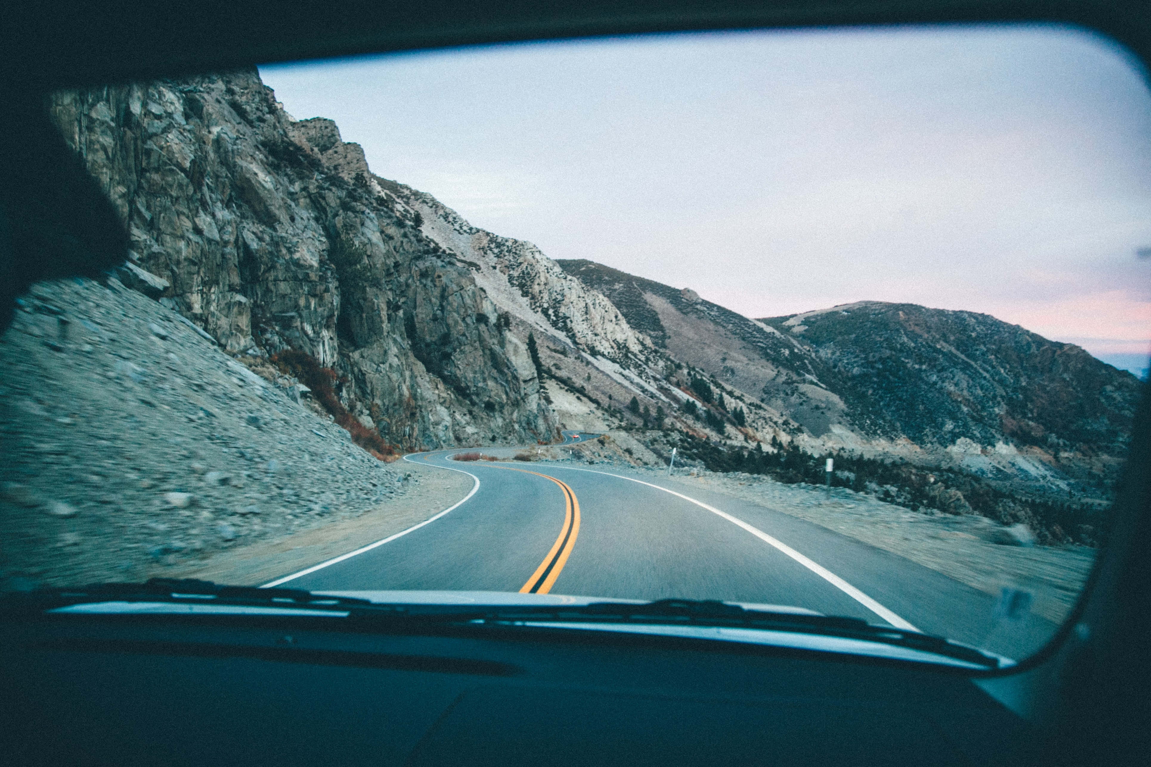 road trip 桌布 壁紙 — 從車內觀看，一條蜿蜒的山路在黃昏時分穿過岩石地形向前延伸。