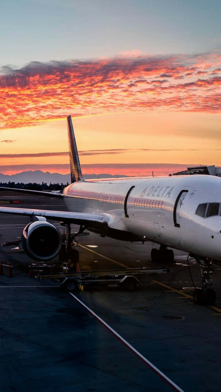從飛機窗戶望見飛機起飛 壁紙 — 一架達美航空飛機停在停機坪上，火紅的橙色和粉紅色雲彩在遠山之上的暮色天空中蔓延。