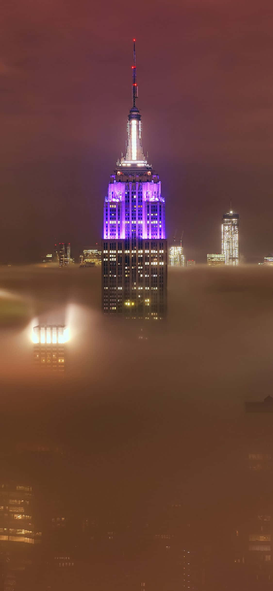 從高樓俯瞰紐約夜景，璀璨奪目 壁紙 — 帝國大廈在紐約市的夜晚透過濃厚的橙色霧氣發出紫色和白色的光芒。