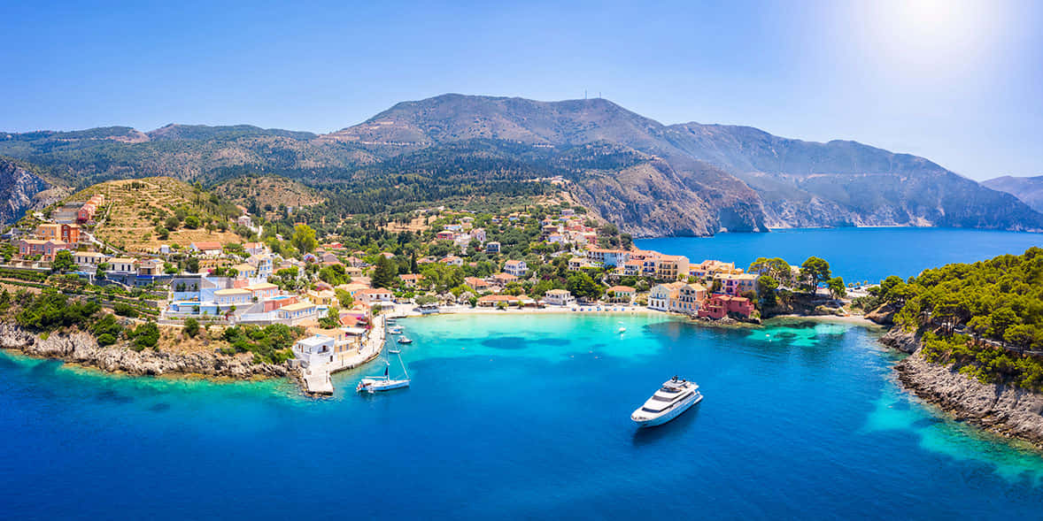 Greek Island 桌布 壁紙 — 從高空俯瞰，一個充滿活力的沿海村莊坐落在碧綠的海水中，背後群山聳立，頭頂是清澈的藍天。