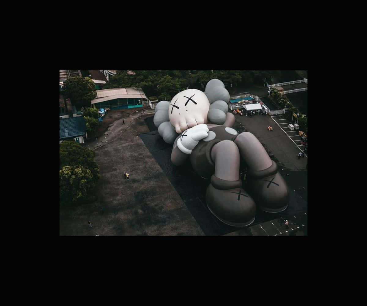 Kaws Holiday 桌布 壁紙 — 從高空俯瞰，一個巨大的KAWS Companion雕塑躺臥在鋪砌區域，四周環繞樹木和小型建築物。