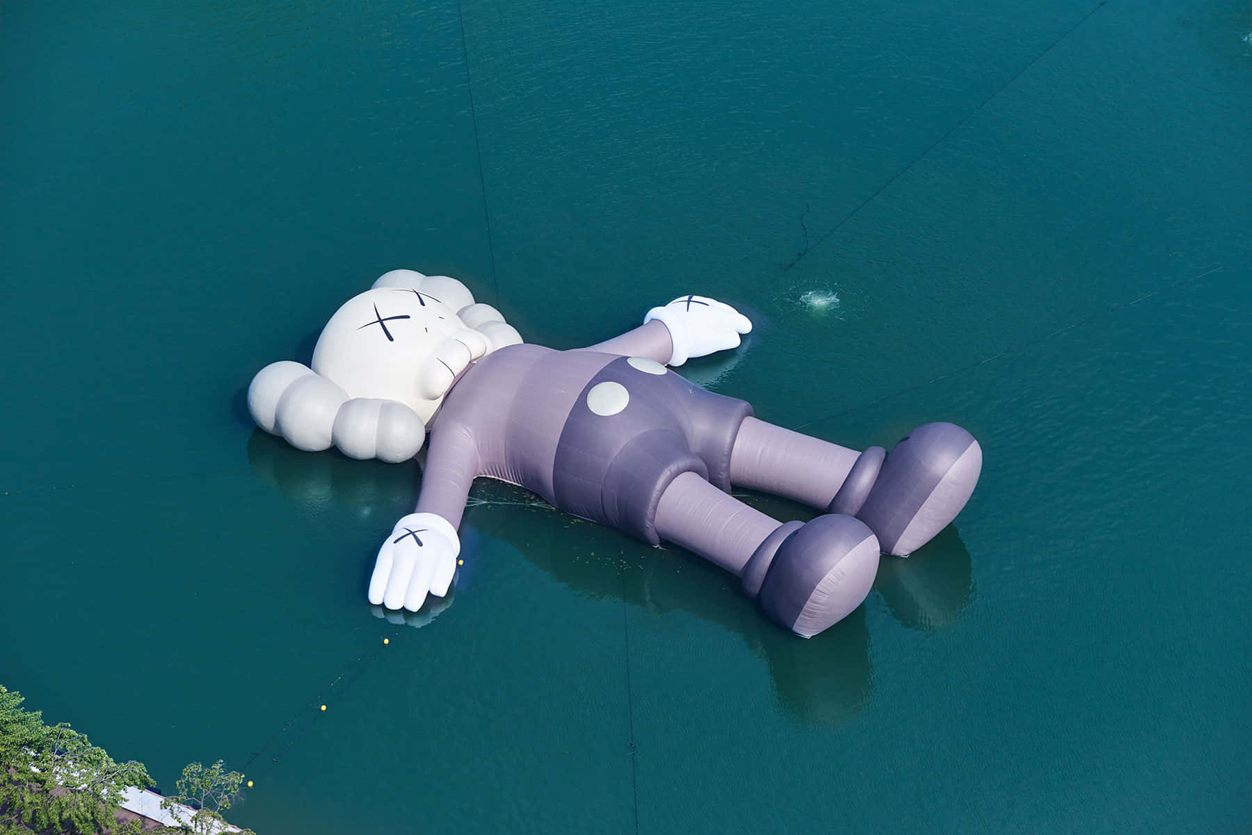 Kaws Holiday 桌布 壁紙 — 從高空俯瞰，一個巨大的充氣KAWS角色平靜地漂浮在寧靜的碧綠水面上。