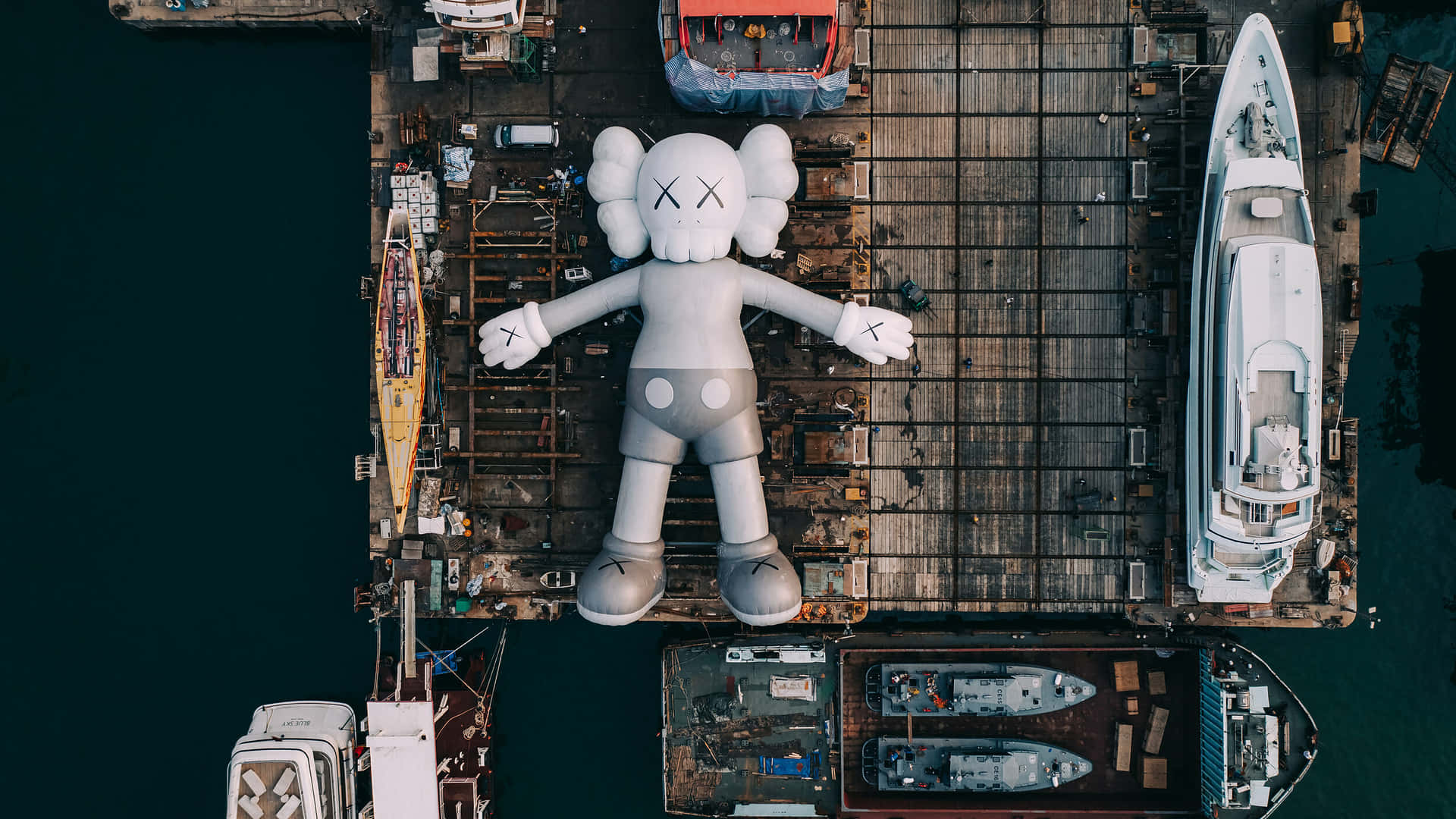 Kaws Holiday 桌布 壁紙 — 從高空俯瞰，一個巨大的白色KAWS雕塑躺臥在風化碼頭上，周圍環繞著船隻和工業設備。