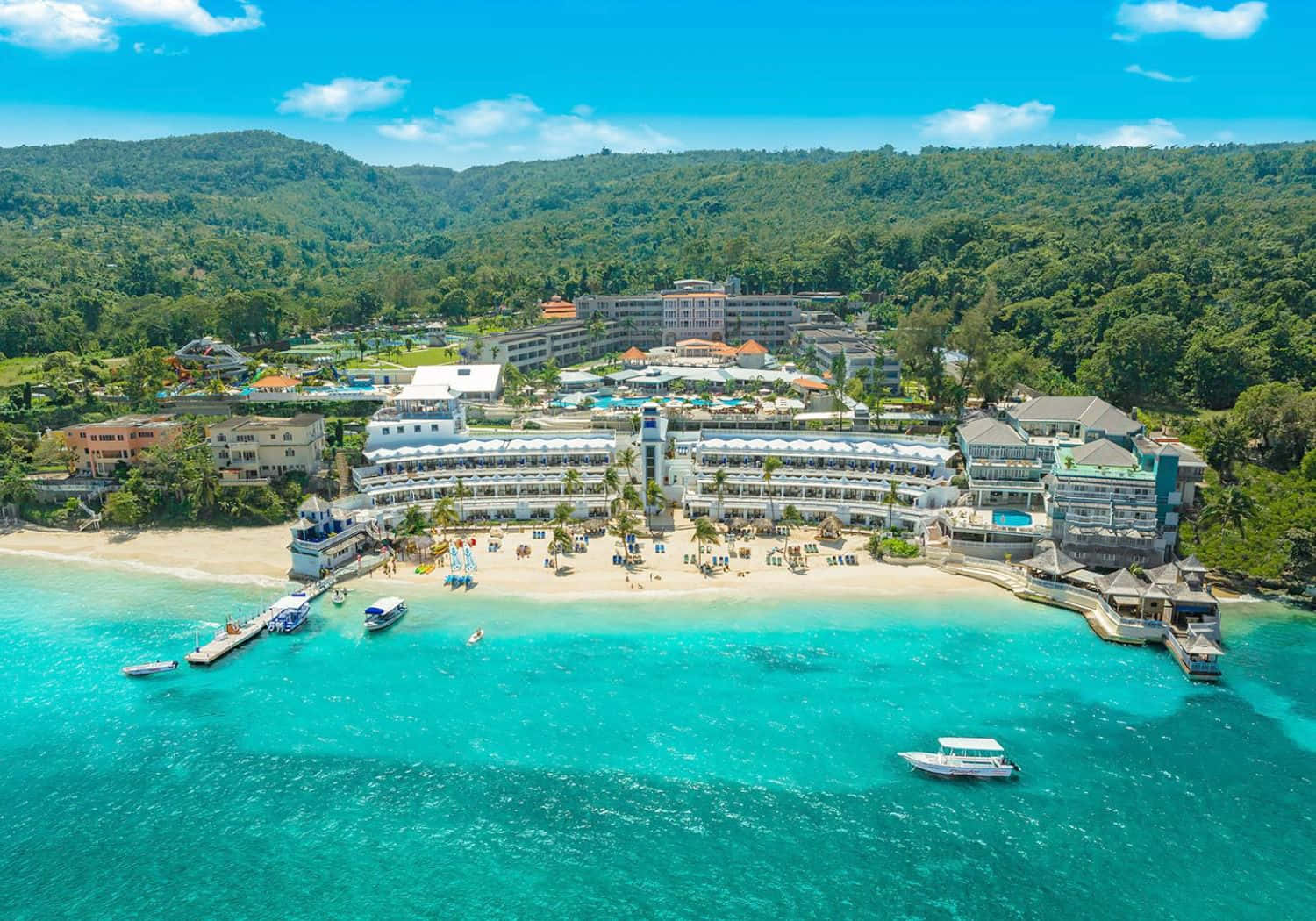Jamaica 桌布 壁紙 — 從高空俯瞰，一個豪華海灘度假村坐落在碧綠海水與翠綠山丘之間，上方是明亮的藍天。